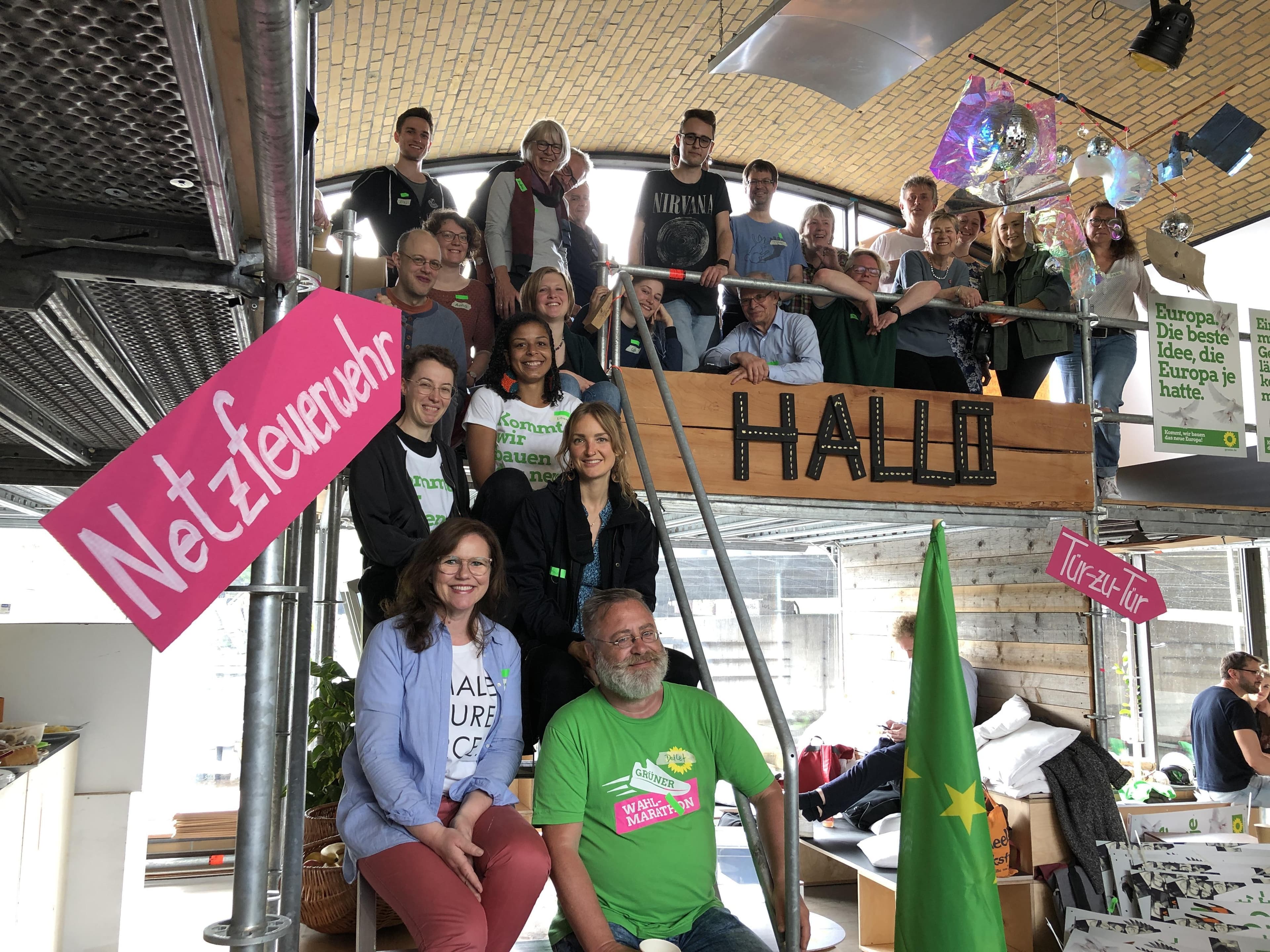 Einige Menschen sitzen auf einer Treppe und stehen am Geländer eine Etage darüber. Auf sie zeigt ein Magenta-Schild mit der Aufschrift "Netzfeuerwehr".