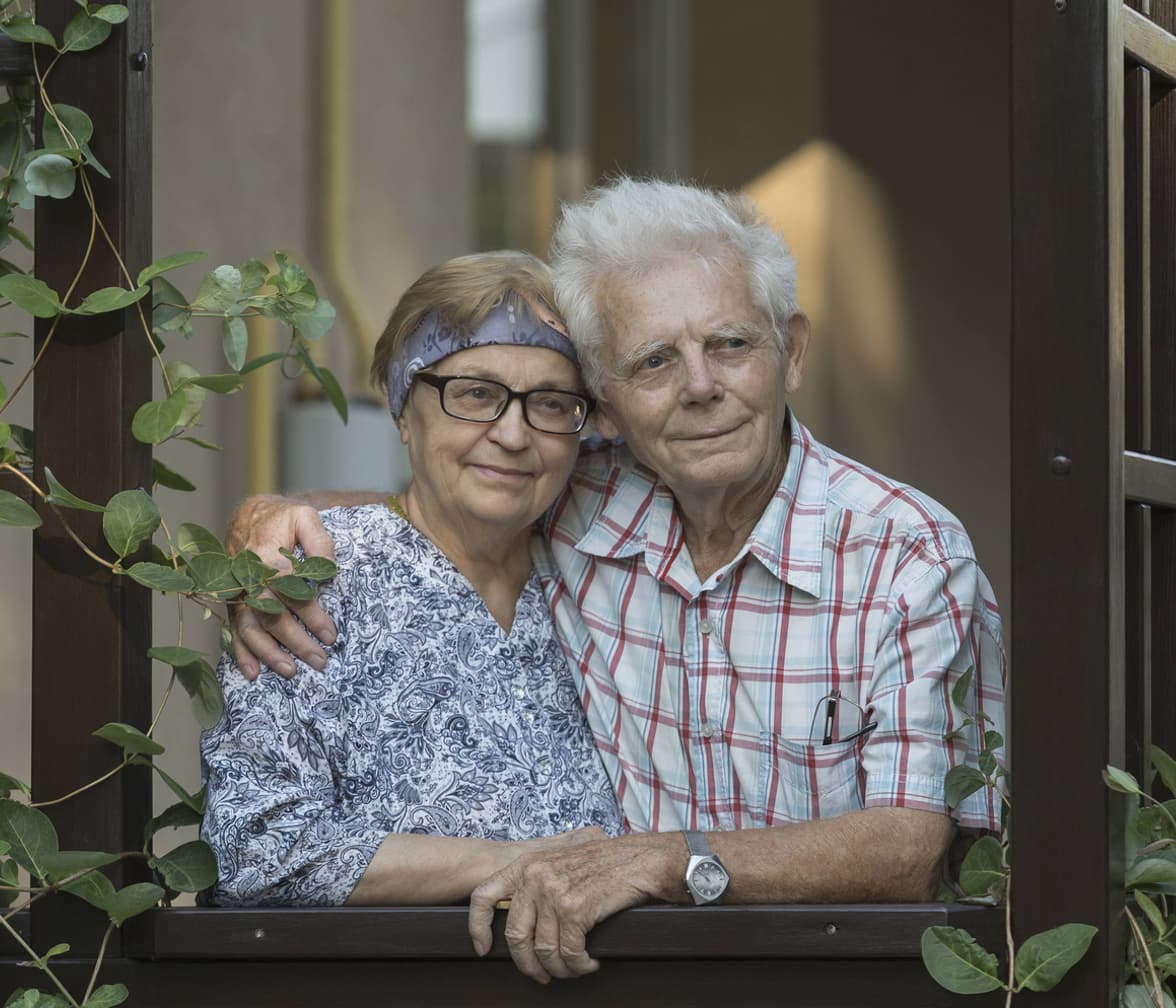 Ein älteres Pärchen steht in liebevoller Umarmung am Fenster und blickt hinaus.