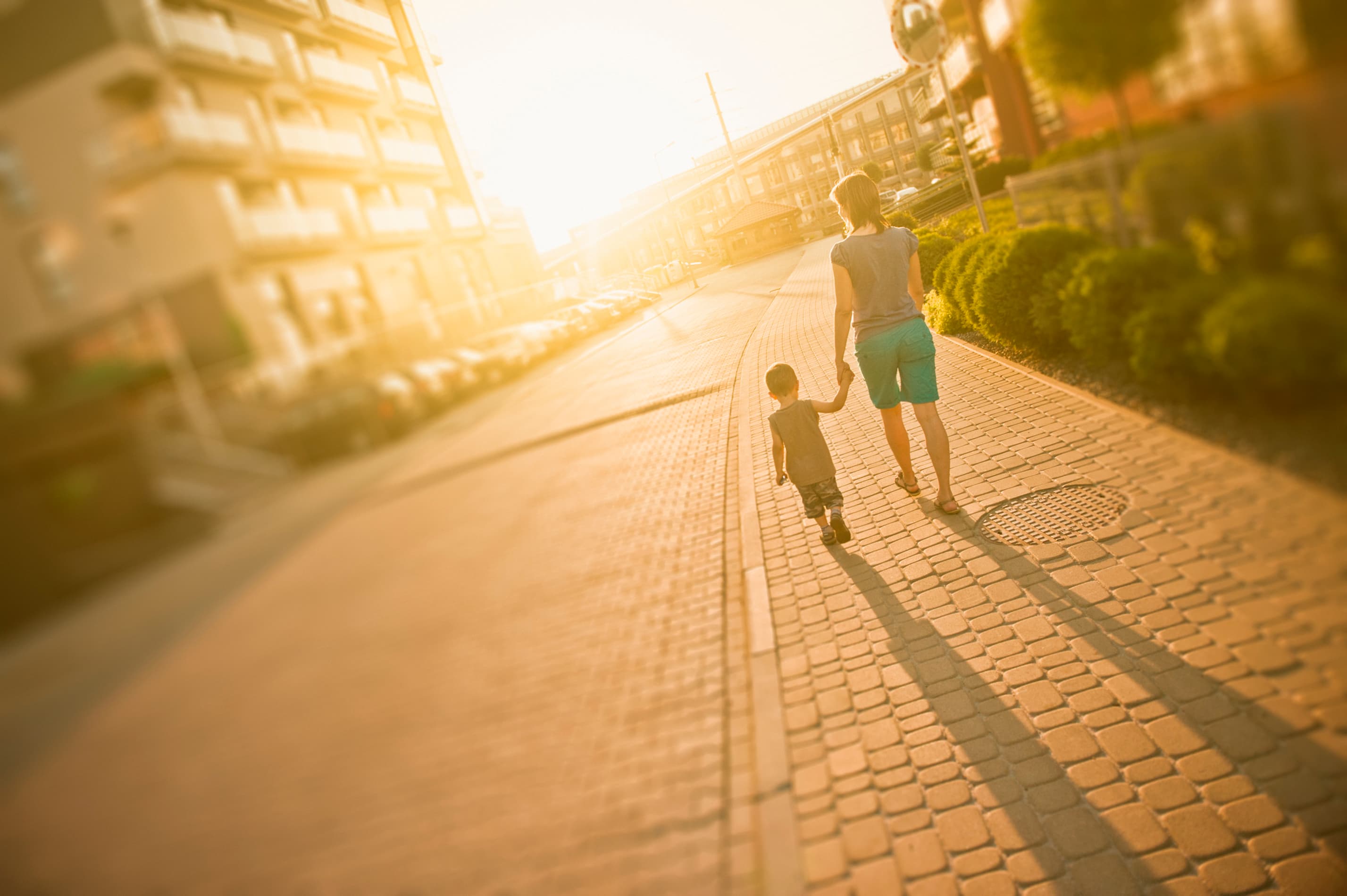 Eine Frau spaziert mit ihrem Sohn Hand in Hand durch ein Wohnviertel. Die Sonne scheint.