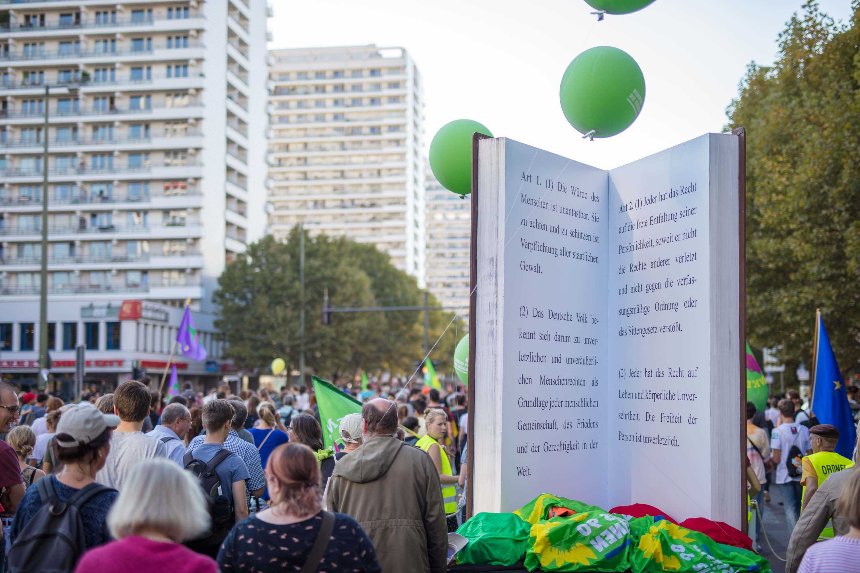 Viele Menschen gehen auf einer Demo durch die Straßen. Die Menge ist von hinten fotografiert. Außerdem ist ein aufgeschlagenes Buch in Übergröße zu erkennen.