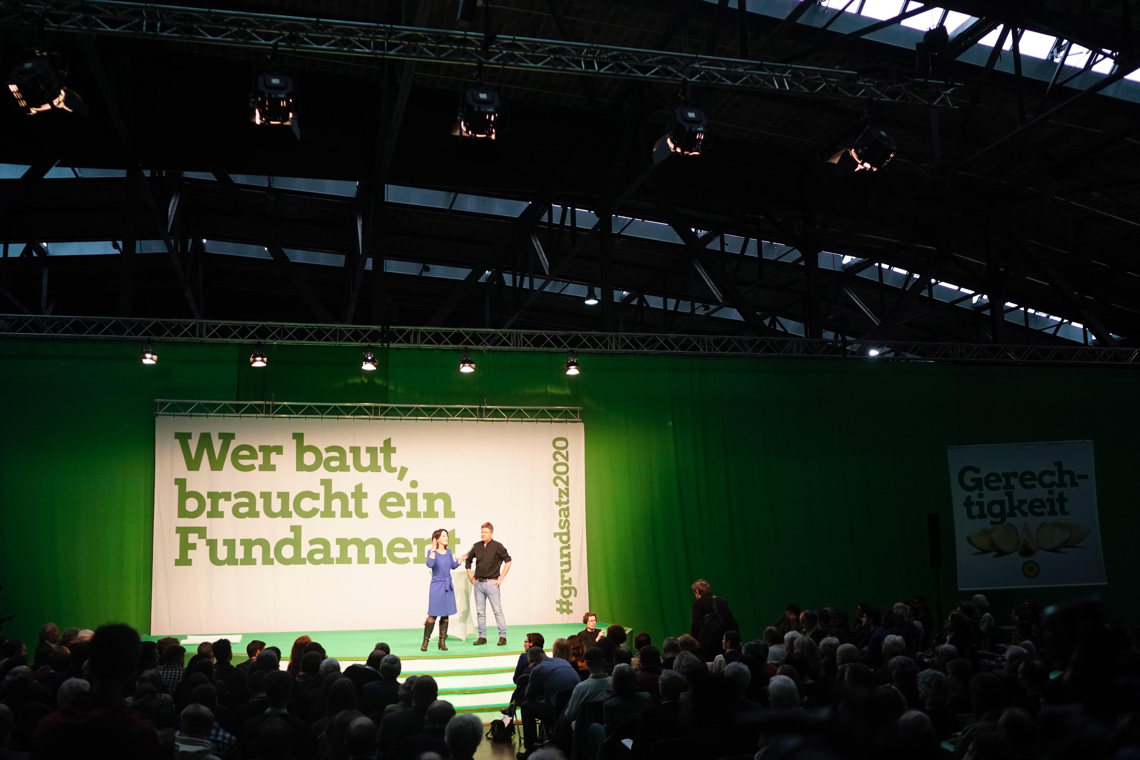 Abgedunkelte Halle mit Beleuchtung auf der Bühne. Zwei Personen stehen vor dem Publikum, im Hintergrund steht: "Wer baut, braucht ein Fundament."