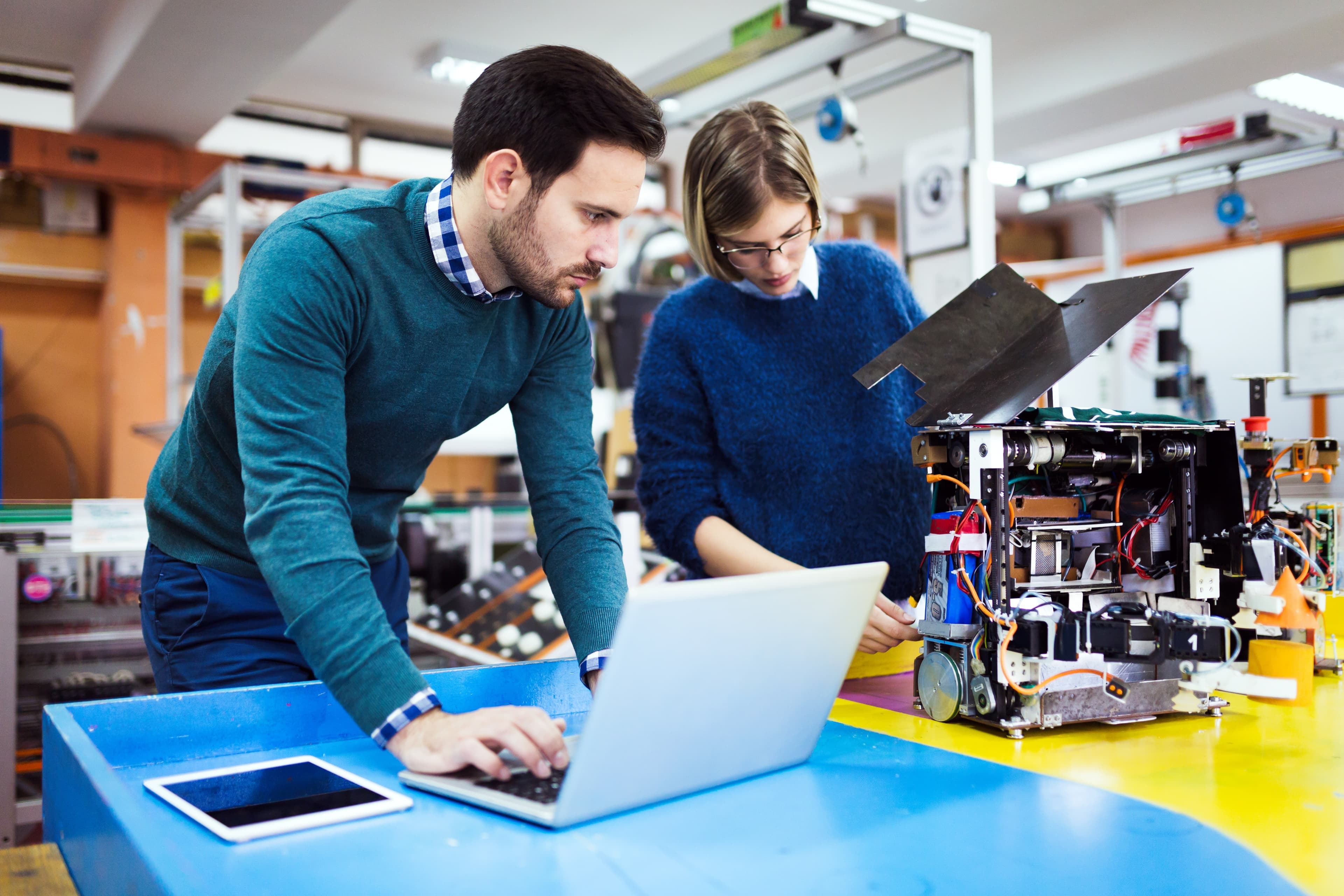 Zwei Personen stehen an einem Tisch und arbeiten. Der Mann ist am Laptop mit dem Blick zu dem Roboter, an dem die Frau arbeitet.