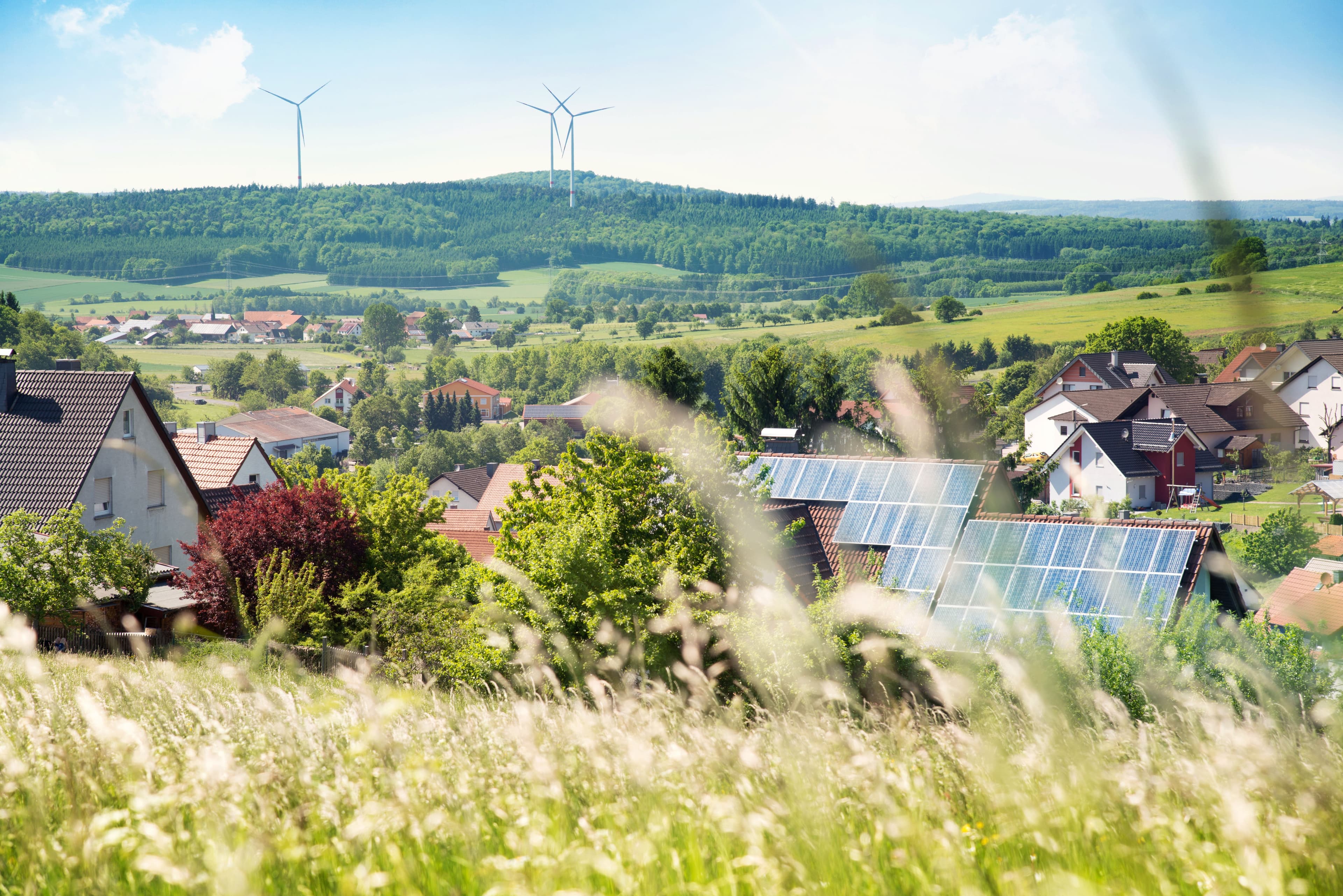 Eine Stadt in grüner Landschaft. Einige Häuser haben Solarzellen auf dem Dach, im Hintergrund sind Windenergieanlagen zu sehen.