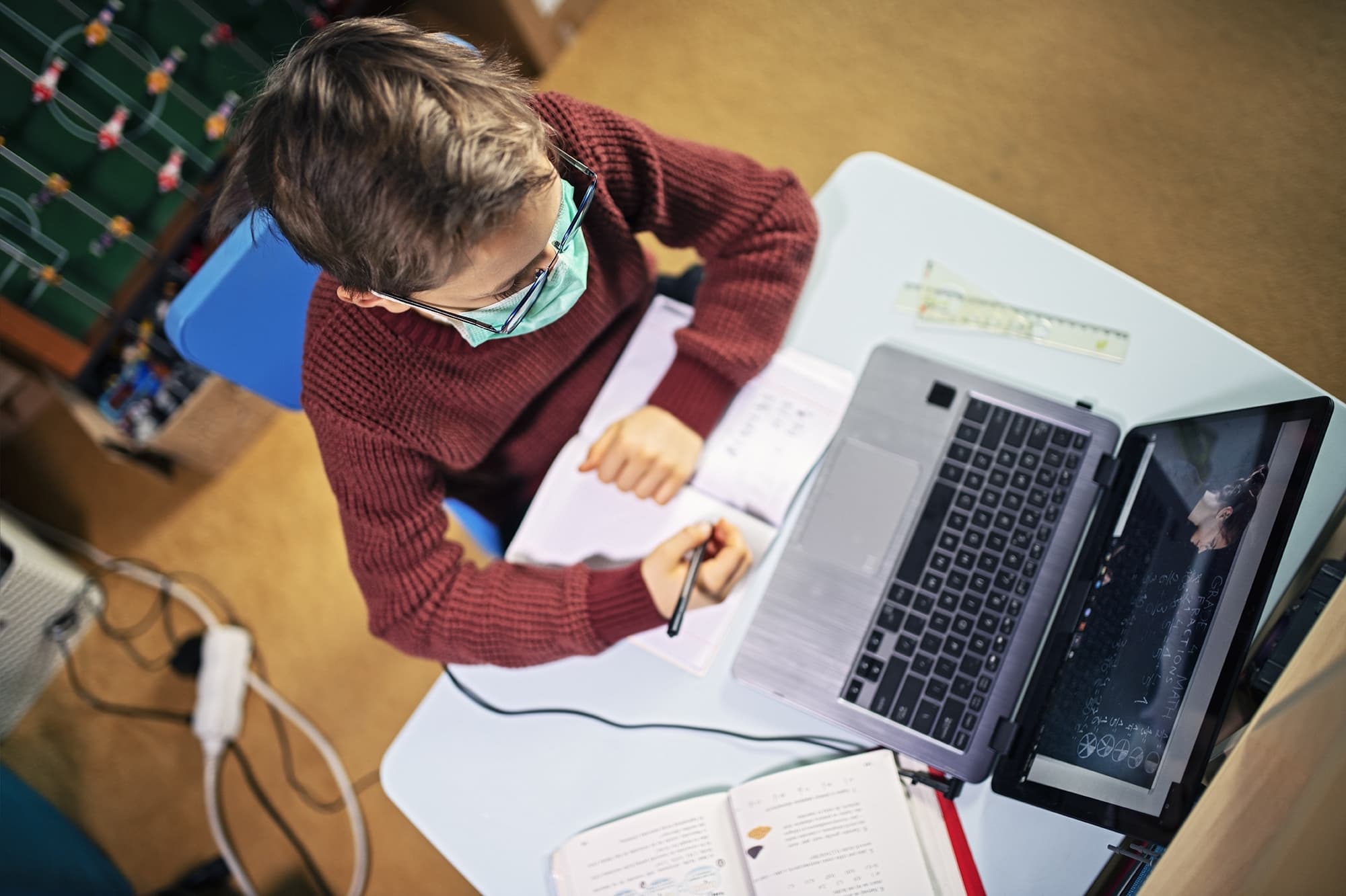 Junge mit Mund-Nase Maske sitzt vor einem Laptop und schreibt in ein Heft.