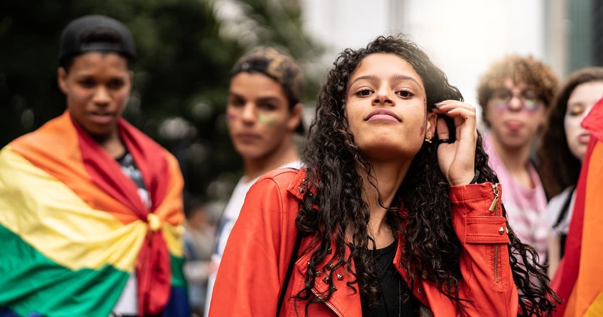 Eine Gruppe Jugendlicher schaut in die Kamera. Im Vordergrund ist ein Mädchen mit dunklen Locken, das selbstbewusst in die Kamera blickt, ein Junge im Hintergrund hat die Pride-Fahne um die Schultern gelegt.