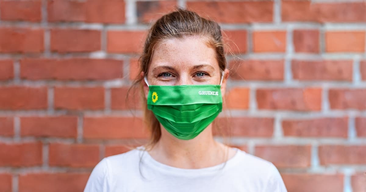 Portait einer blonden jungen Frau mit grüner Maske vor einer Backsteinwand