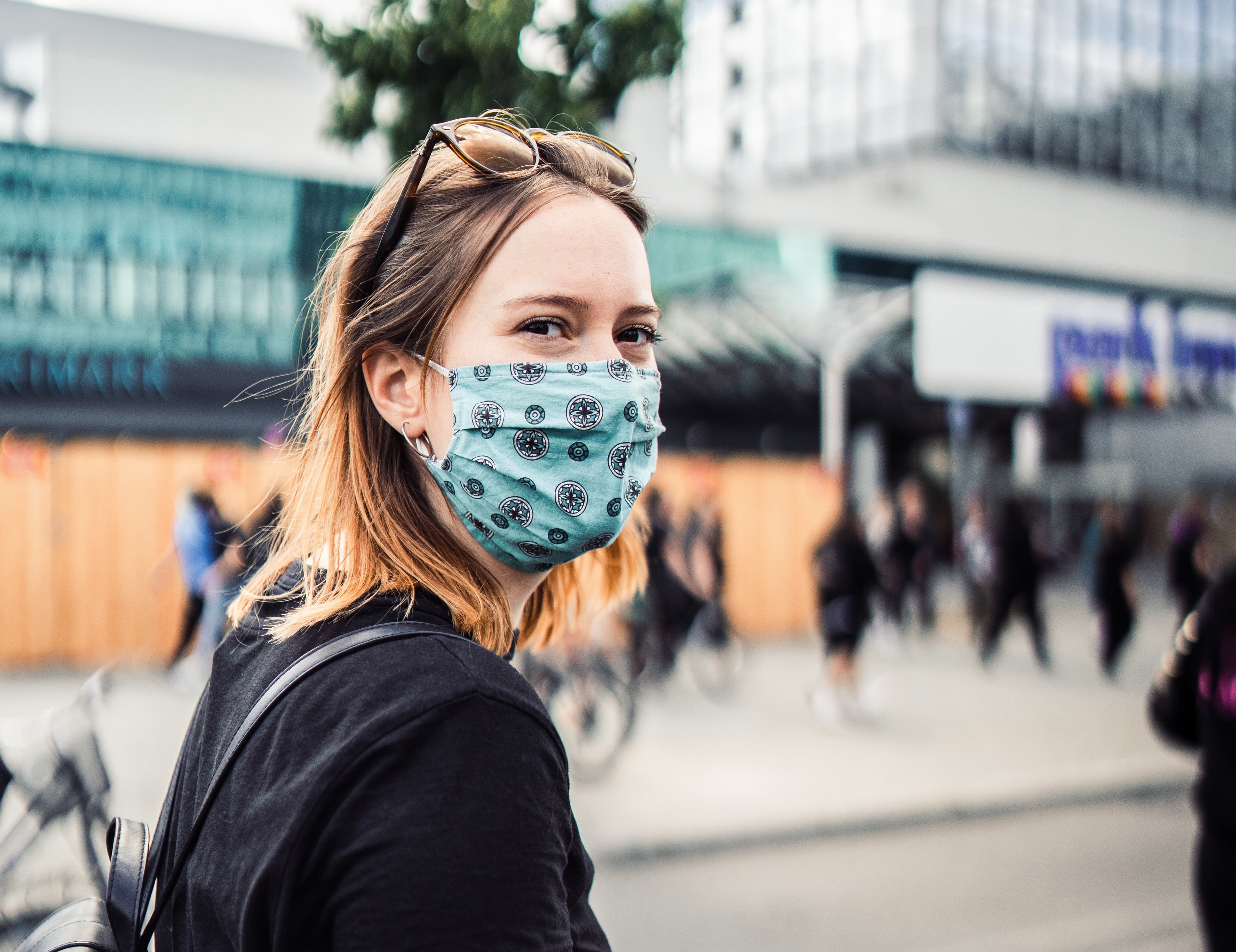 Portrait einer jungen blonden Frau mit Sonnebrille in den Haaren und türkis gemustertem Mund-Nase-Schutz. Im HIntergrund sieht man eine U-Bahn-Station.