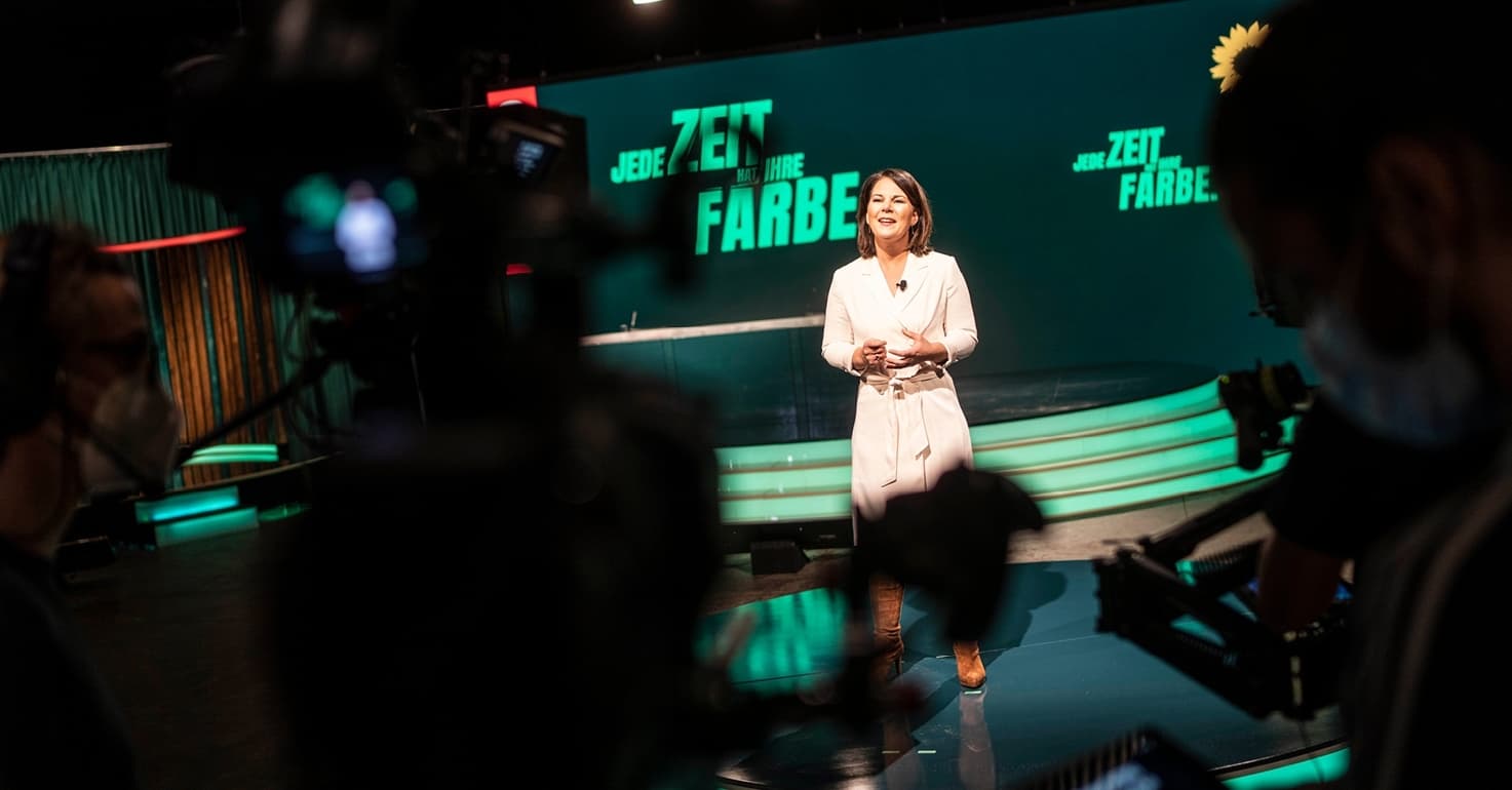 Annalena Baerbock steht vor einer Bühne und einer Grünen Wand mit der AUfschrift: "Jede Zeit hat ihre Farbe". Im Vordergrund sieht man eine Kamera.