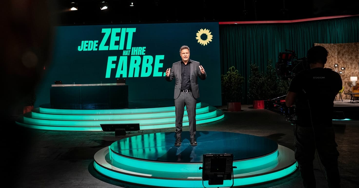 Robert Habeck steht auf einer runden Bühne mit grünen Lichtern am Sockel. Hinter ihm eine Wand mit einer Grünen Aufschrift: Jede Zeit hat ihre Farbe" Im Vorderungrund sieht man im Dunkeln einen Kameramann.