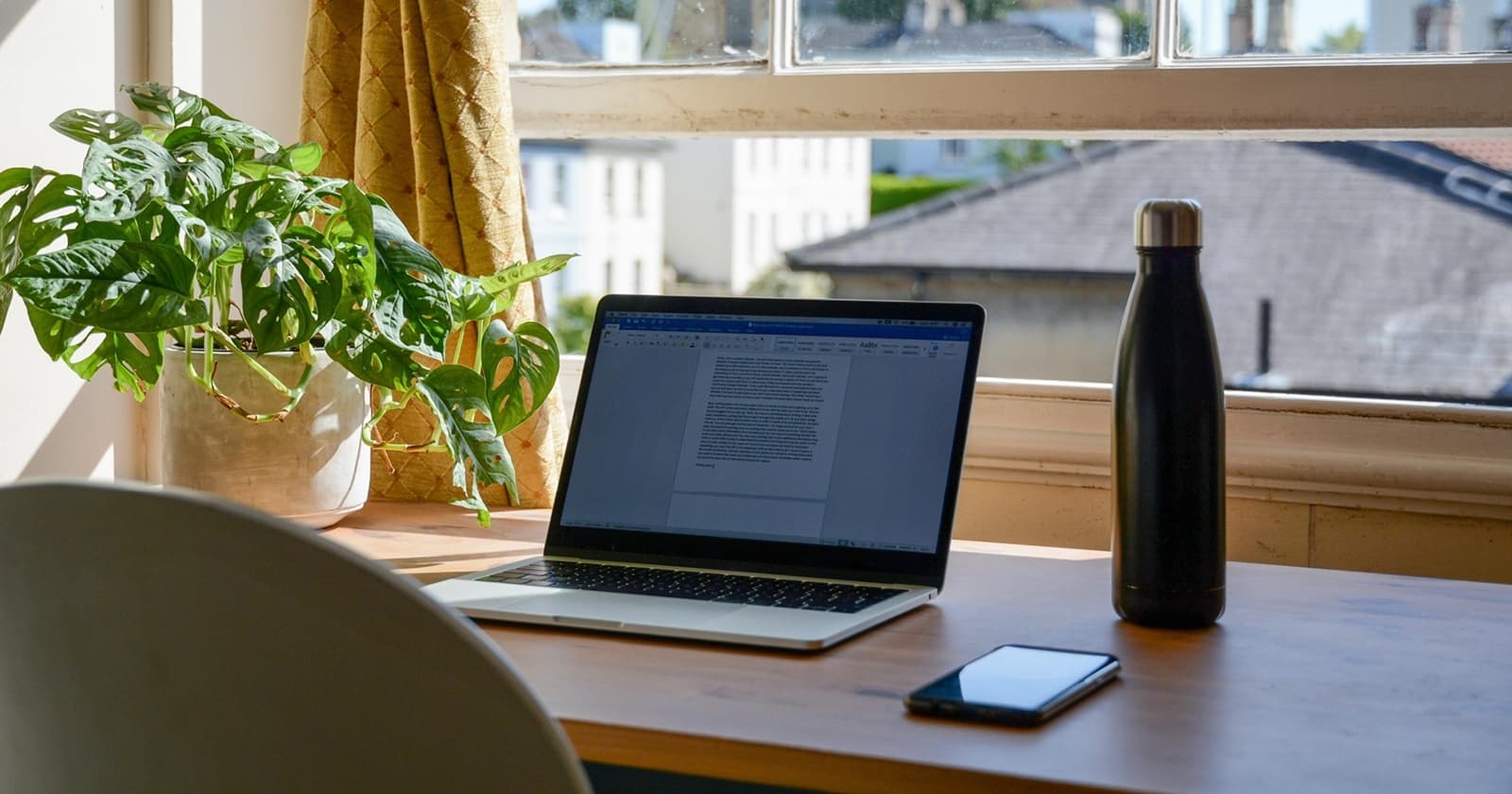 Laptop steht aufgeklappt auf einem hölzernen Schreibtisch, rechts daneben eine Trinkflasche, links eine Pflanze. Der Blick richtet sich Richtung Fenster wo man Dächer sieht.