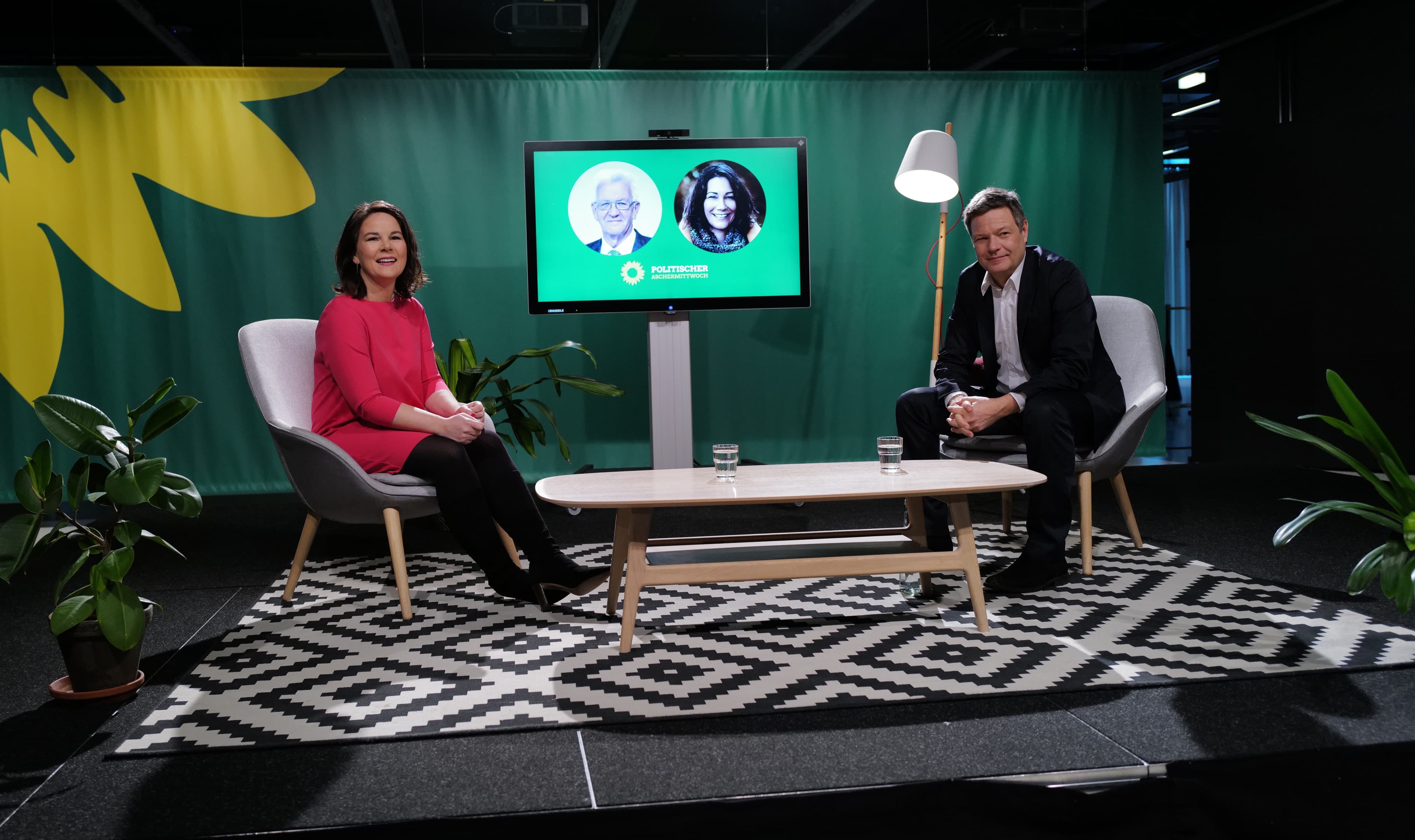 Annalena Baerbock (links) und Robert Habeck sitzen auf einer Bühne mit einem schwarz-weiß gemusterten Teppich und einem Beistelltisch. Rechts und links stehen Kübelpflanzen, hinter den beiden ist ein Bildschirm, auf dem Winfried Kretschmann und Anne Spiegel zu sehen sind.