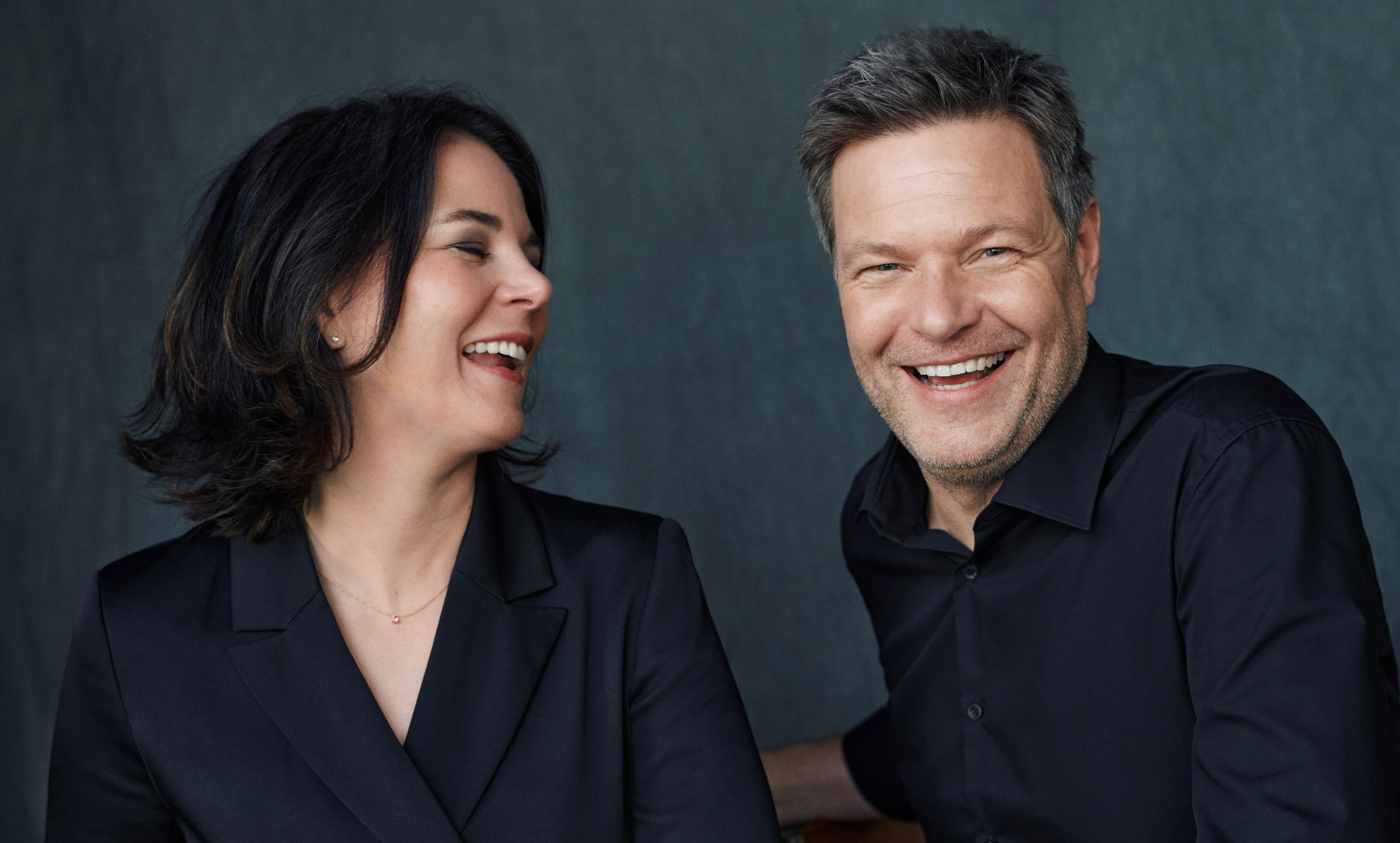 Annalena Baerbock (links) und Robert Habeck (rechts) sitzen nebeinander und lachen. Sie tragen beide dunkle Oberbekleidung und sind bis zum Torso zu sehen. Annalena Baerbock sieht zu Robert Habeck.