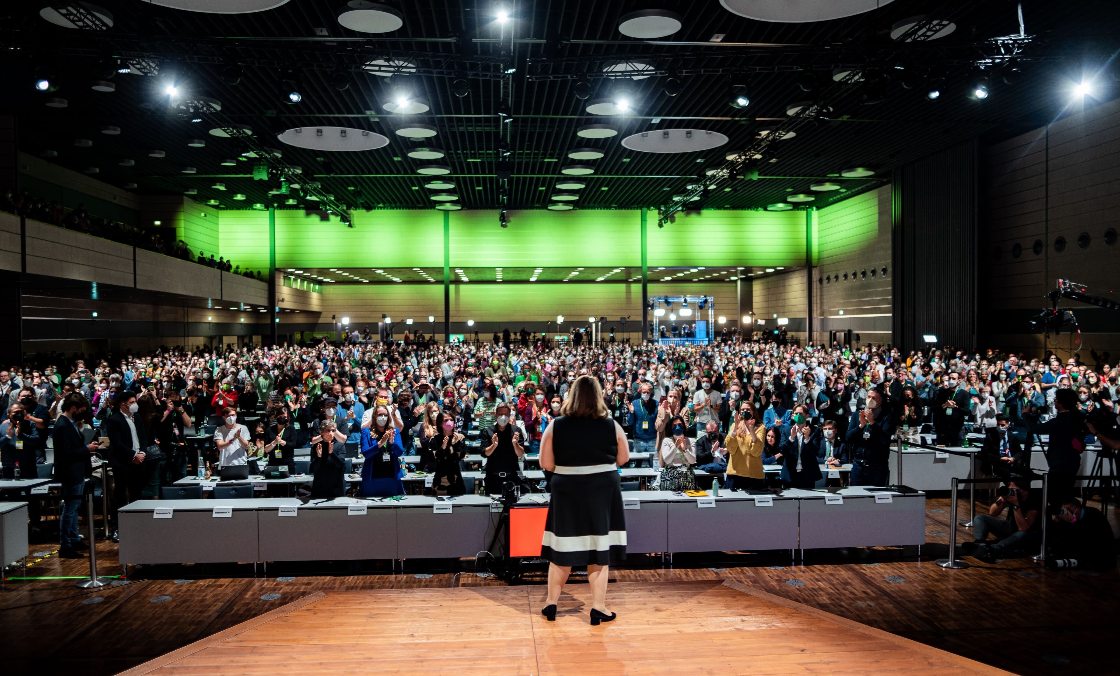 Ricarda Lang steht in einem schwarzen Kleid mit dem Rücken zur Kamera auf der Bühne und schaut auf eine Halle voll mit Tischen und Menschen, die stehen und klatschen.