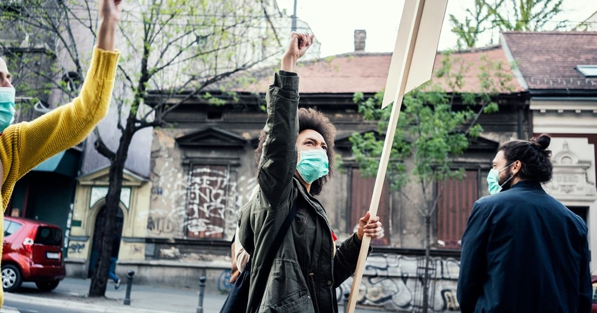 EMan sieht Demonstrant*innen mit erhobener Faust, Mund-Nasen-Schutz und Sicherheitsabstand. Im Fokus ist eine Frau, die ein Schild trägt, von dem man nur die Rückseite sieht.