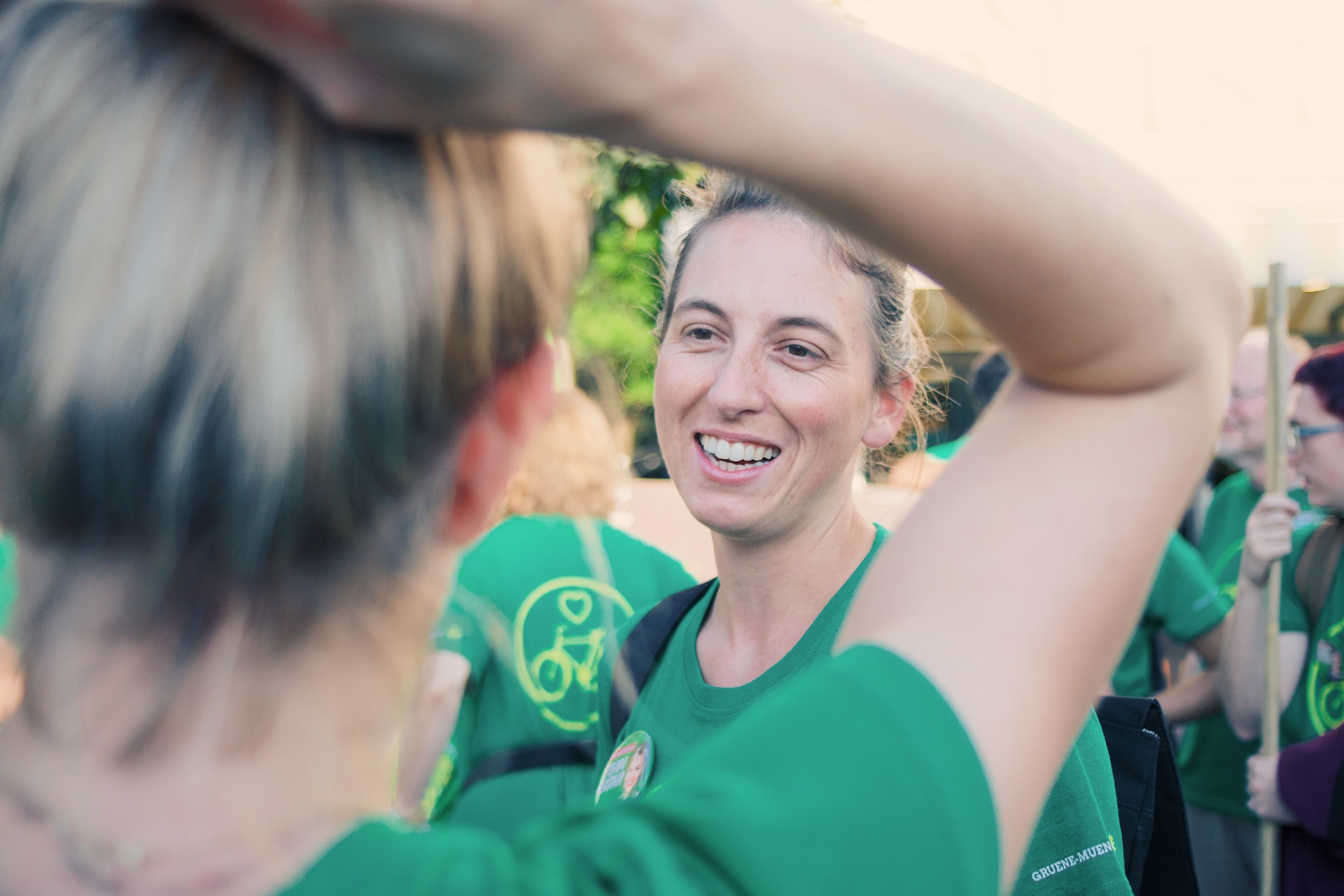 Vordergrund: Zwei Frauen in grünem T-Shirt. Eine mit dem Rücken zur Kamera, die andere frontal. Im Hintergrund befinden sich weitere Personen.