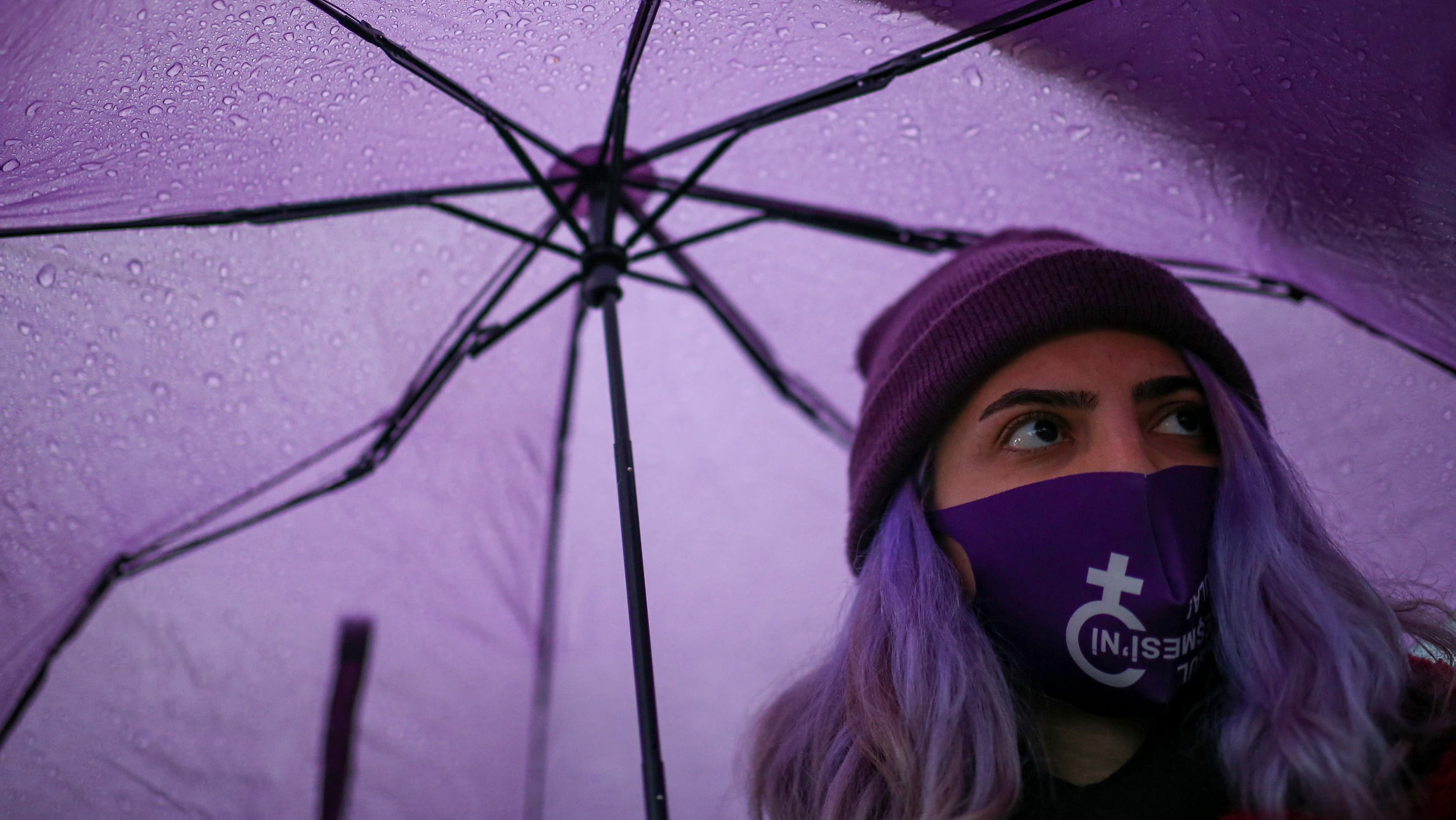 Nahhaufnahme: Eine junge Frau mit violett gefärbten Haaren, einer violetten Mütze und violettem Mund-Nasen-Schutz mit dem Weiblichkeitssymbol darauf unter einem violetten Regenschirm