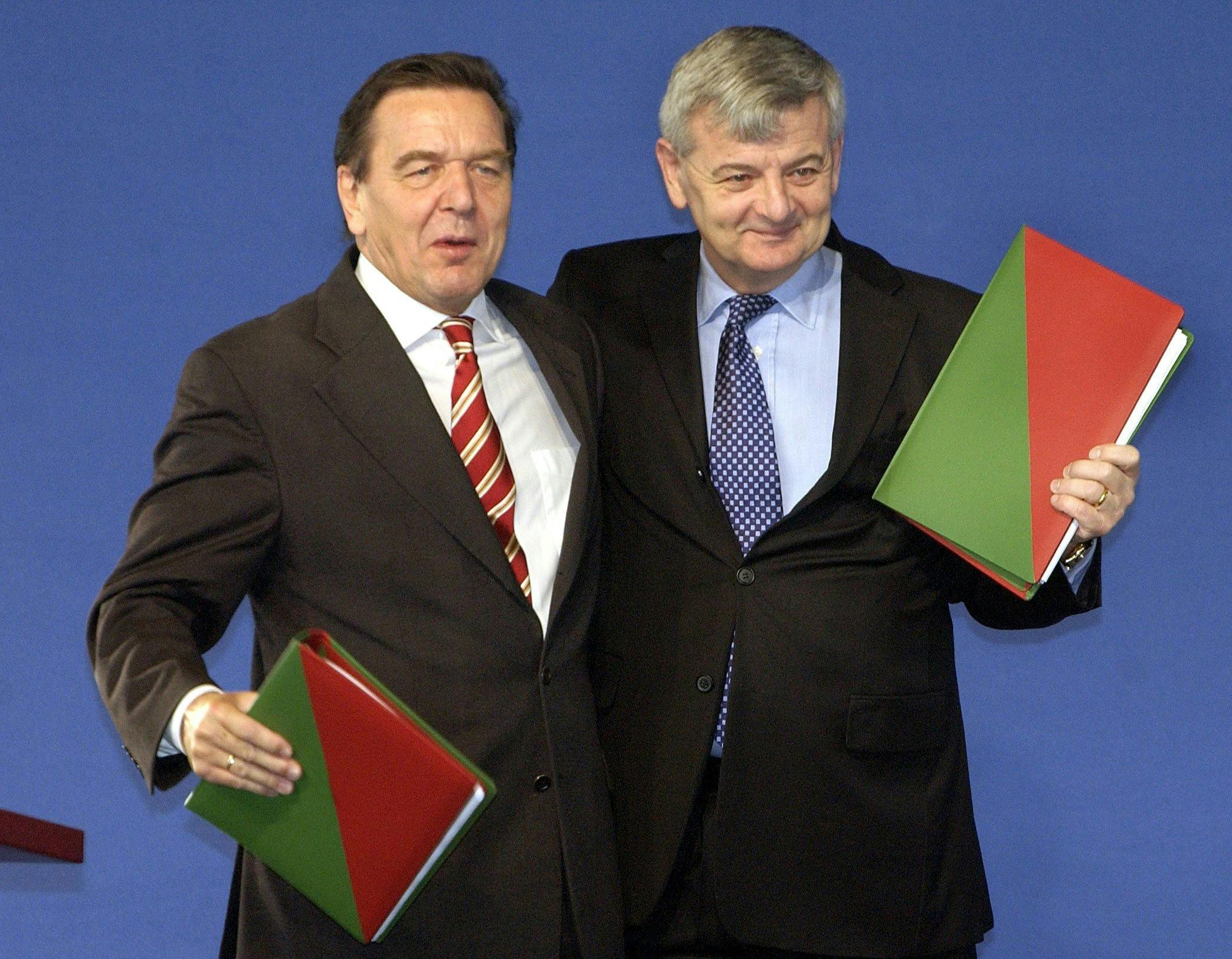 Gerhard Schröder und Joschka Fischer stehen zusammen vor blauem Hintergrund und halten jeweils ein Buch mit grün-rotem Umschlag hoch.