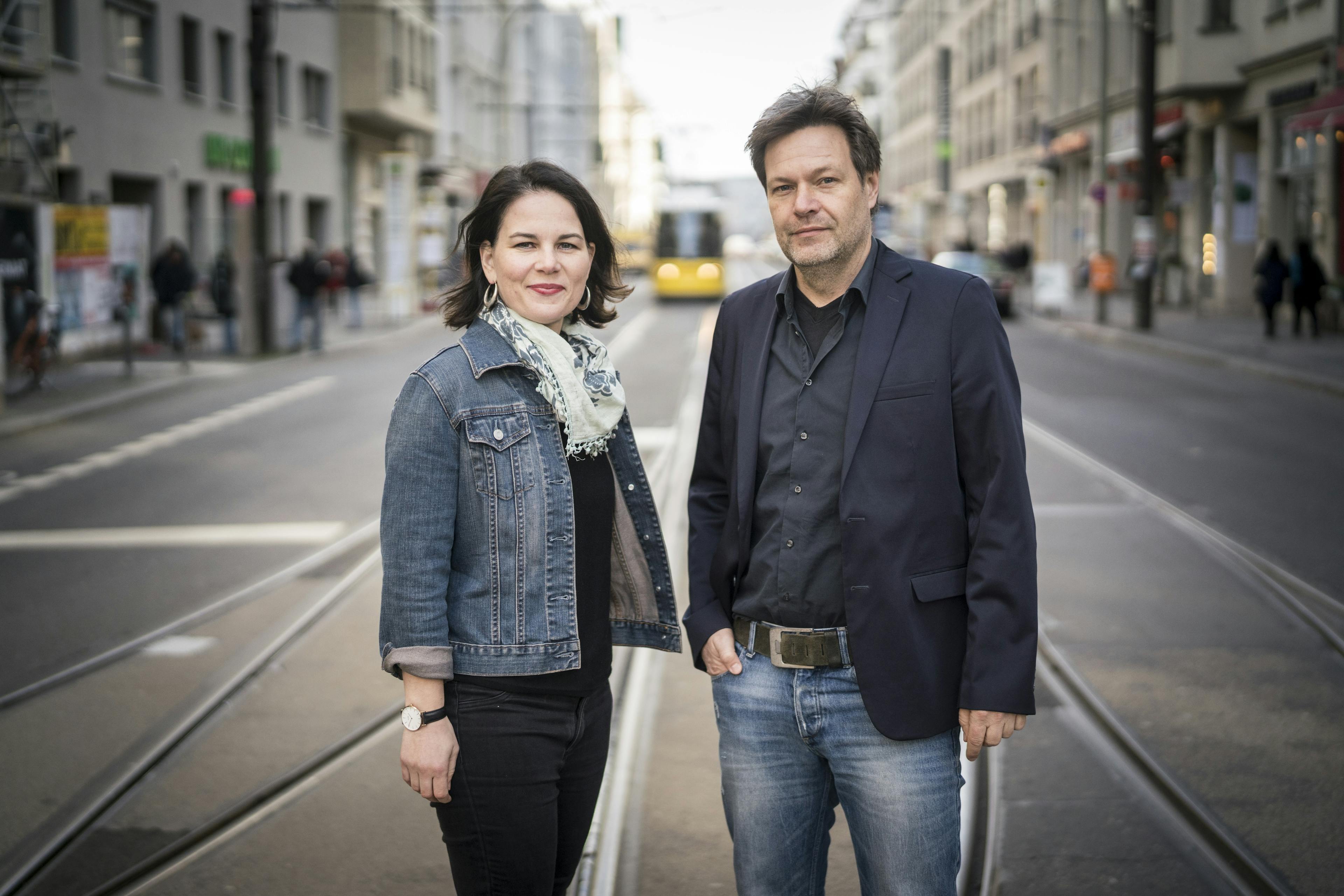 Annalena Baerbock und Robert Habeck stehen gemeinsam mitten auf einer Straße mit Bahnschienen. Im Hintergrund sind Häuser und eine Straßenbahn zu sehen.