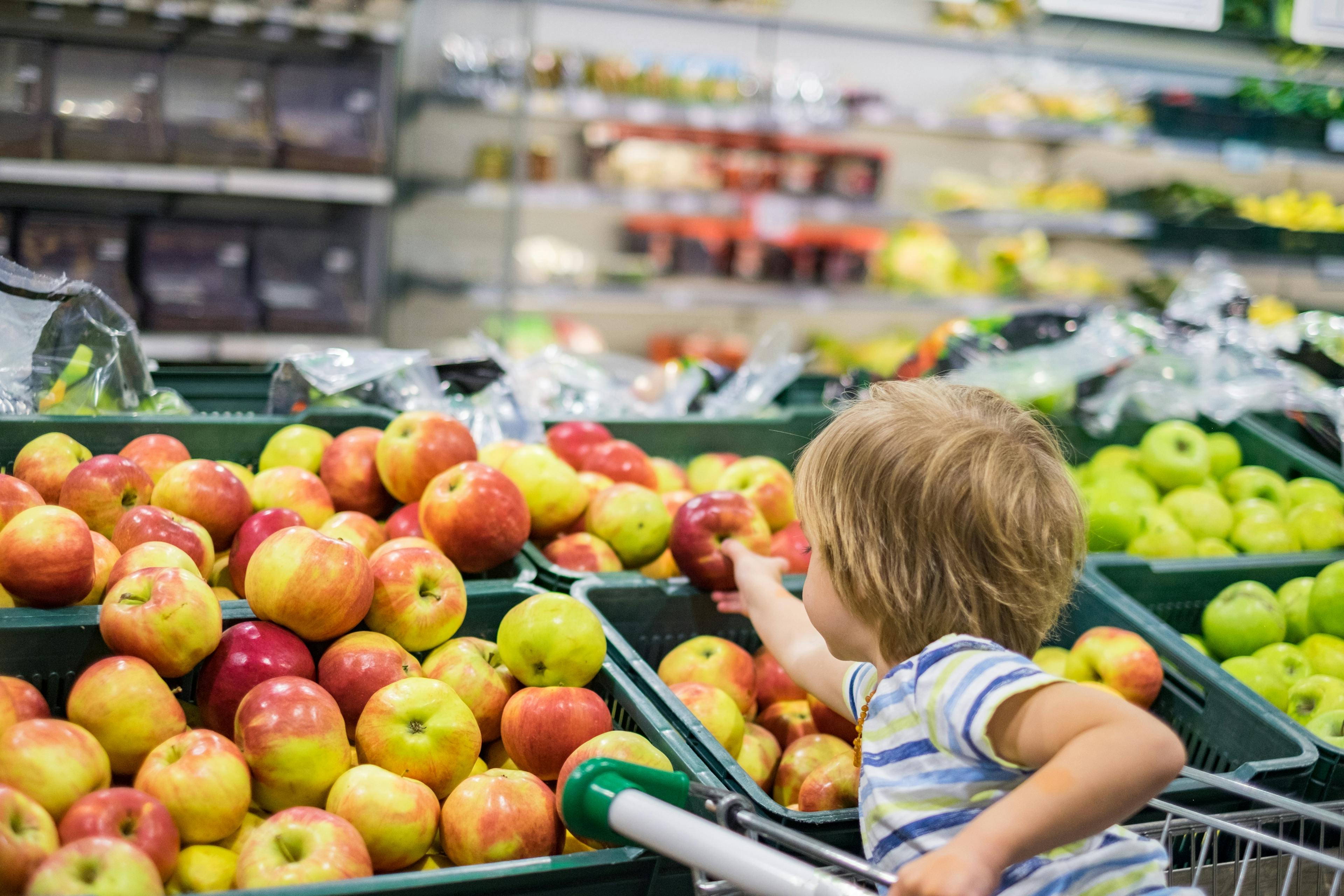 Ein kleines Kind sitzt in einem Einkaufswagen und greift nach einem Apfel aus der Supermarkt-Theke.