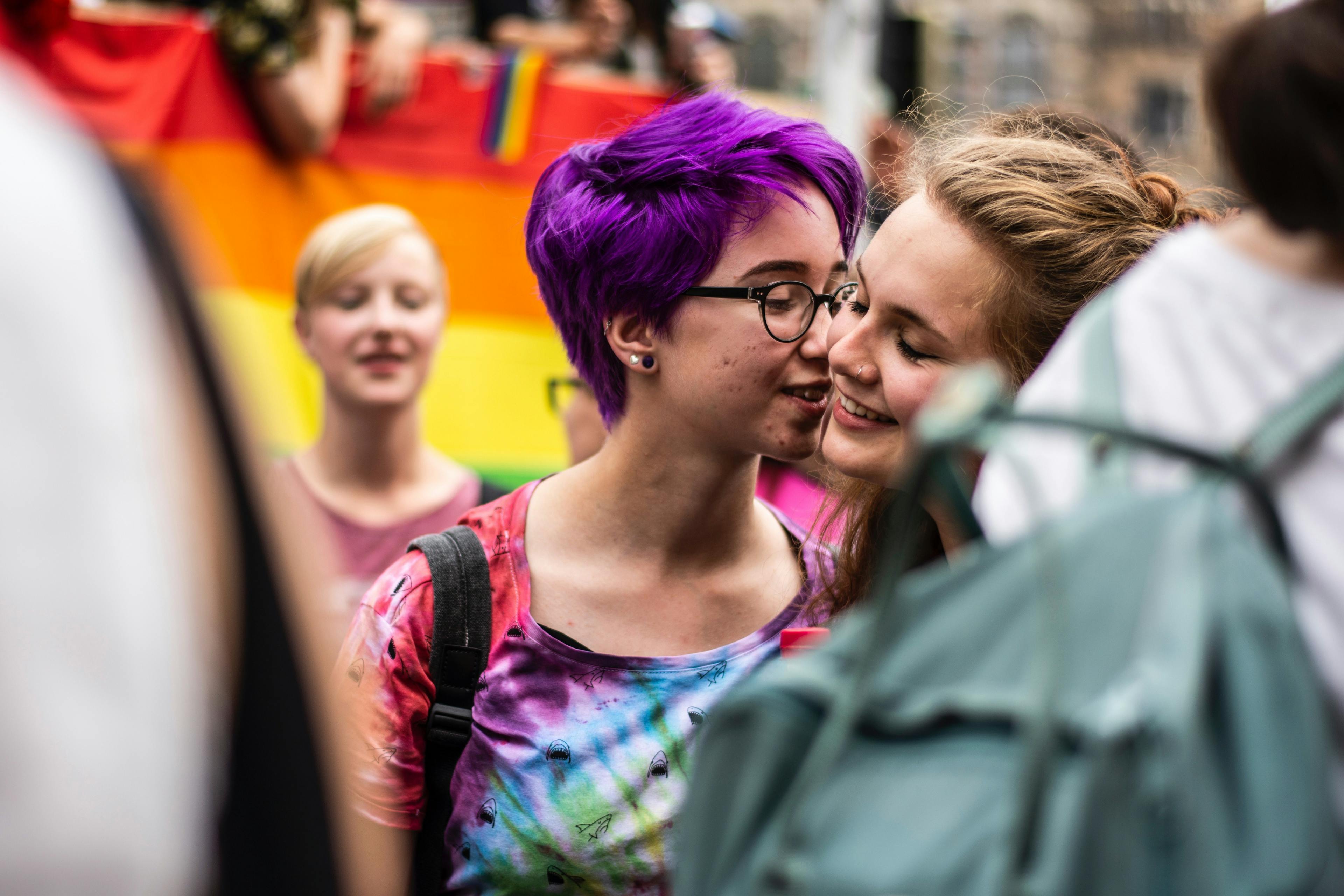 CSD Demo: Zwei junge Frauen lächeln, die eine gibt der anderen einen Kuss auf die Wange.