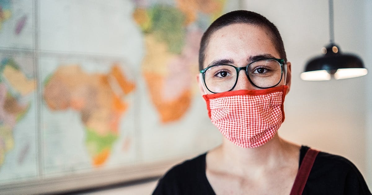 Portrait einer jungen Frau mit rot-weiß karierter Maske, die vor einer Weltkarte steht