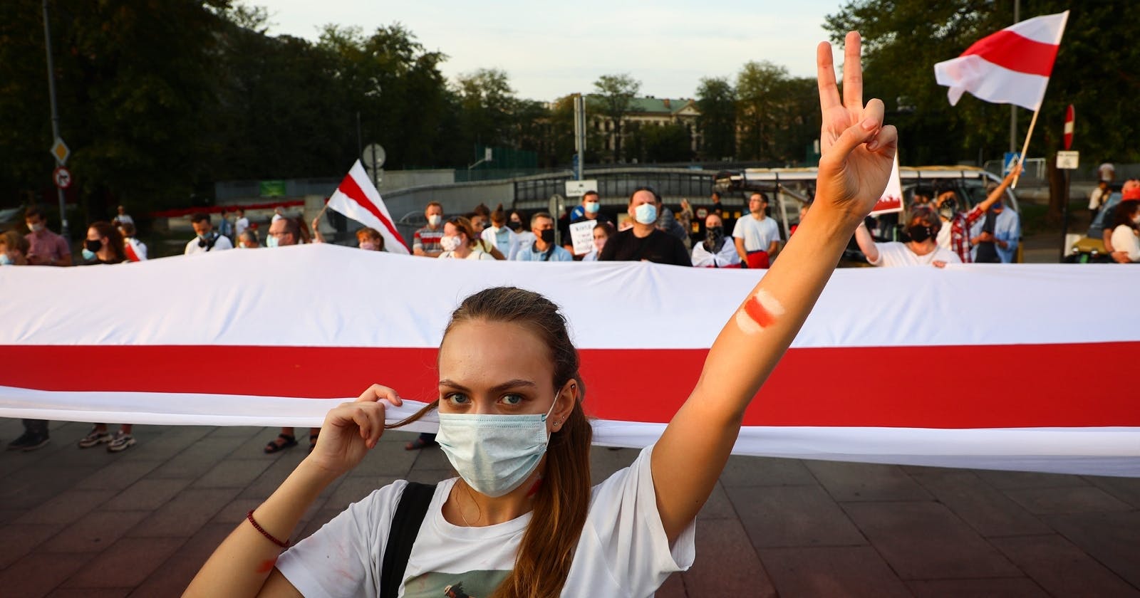 Eine junge Frau im weißen T-Shirt steht vor einer Gruppe von Leuten, die einen riesigen Banner in den belarusischen Nationalfarben vor sich herträgt. Sie reckt den linken Arm hoch, mit der Hand formt sie das "Peace"-Zeichen