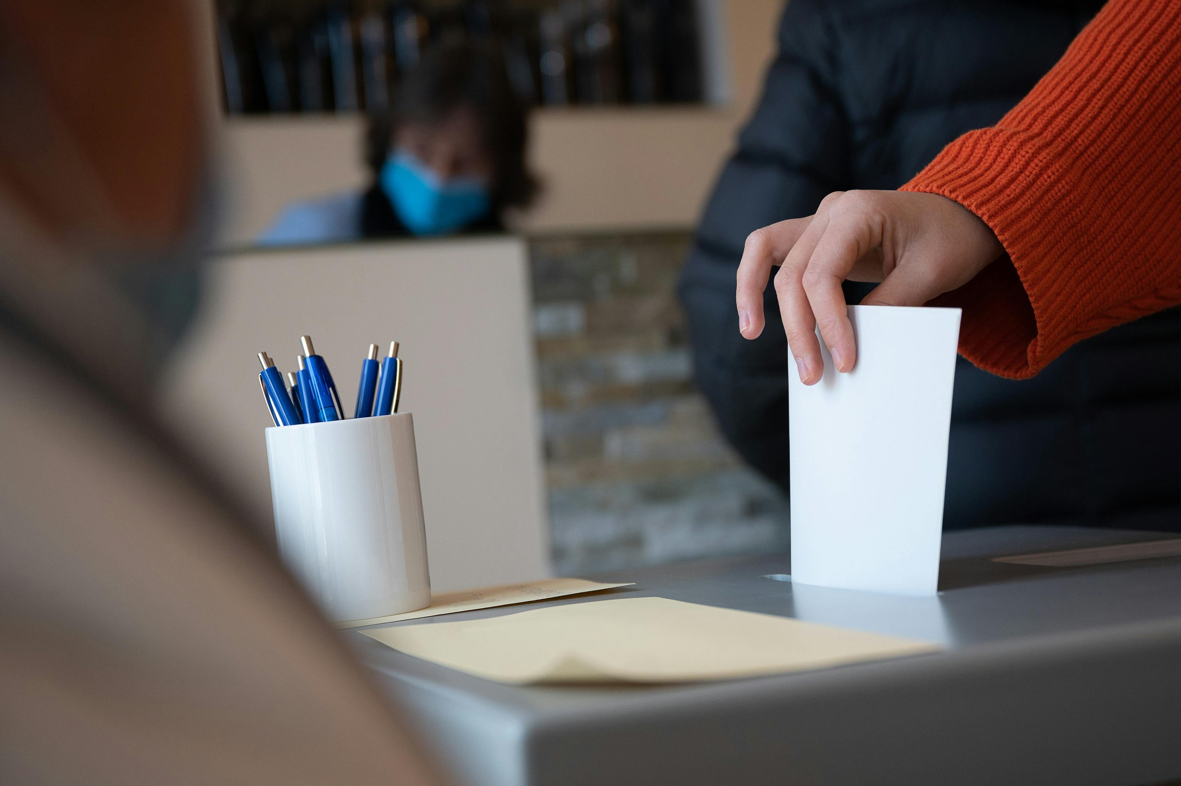 Man sieht eine Wahlurne, in die gerade von einer Person mit rotem Pulli ein Zettel eingeworfen wird.