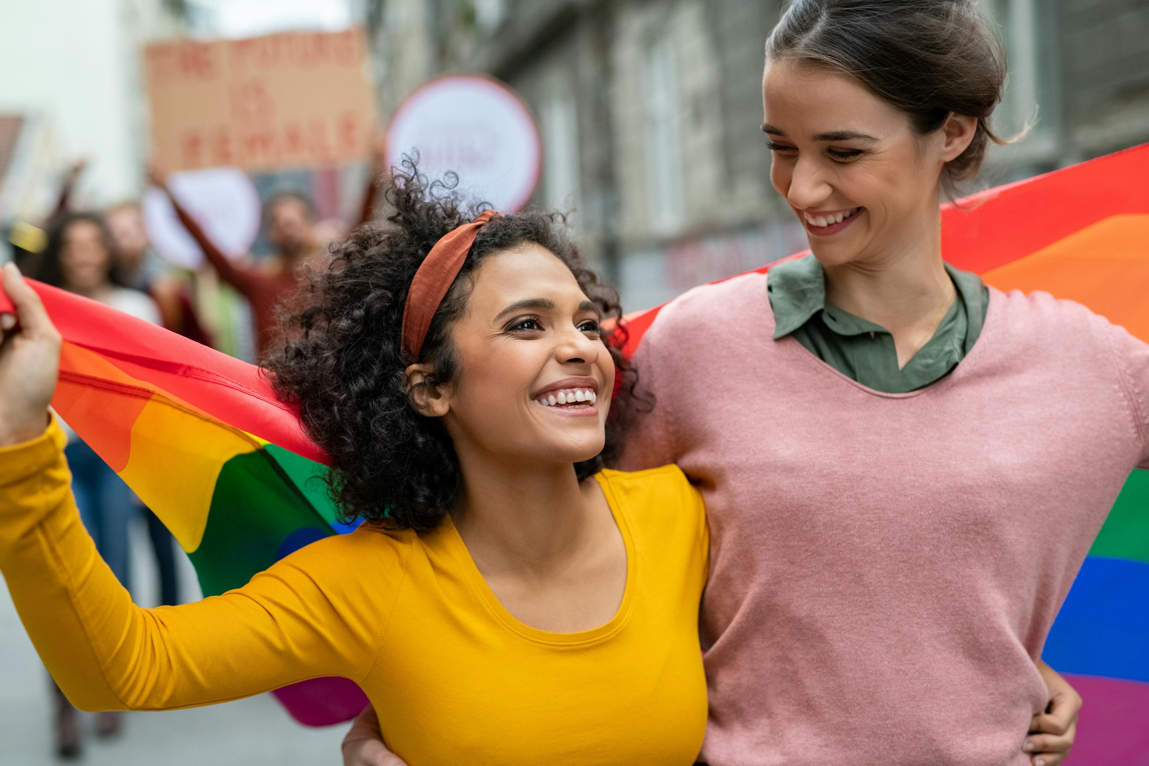 Zwei junge Frauen gehen Arm in Arm und tragen gemeinsam die Pride-Flagge