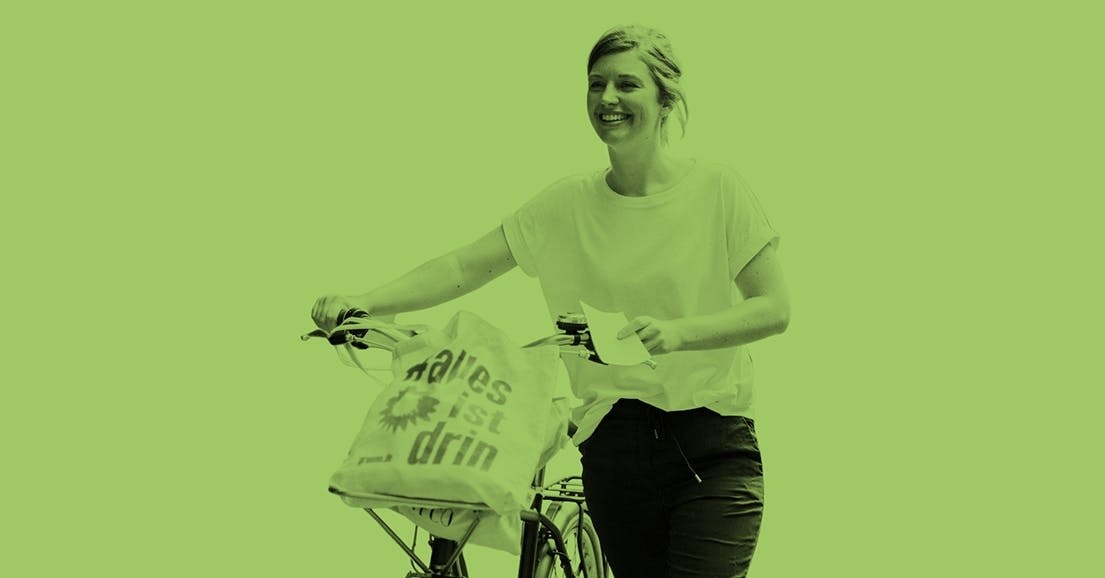 Junge Frau schiebt Fahrrad, im Fahrradkorb liegt ein Jutebeutel mit der Aufschrift: "alles ist drin"