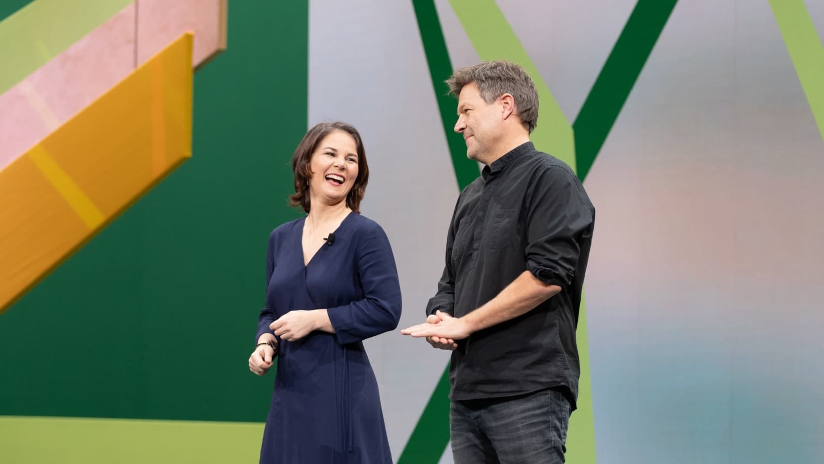 Annalena Baerbock und Robert Habeck stehen nebeneinander vor einem grün-rosanen Hintergrund und lächeln sich an