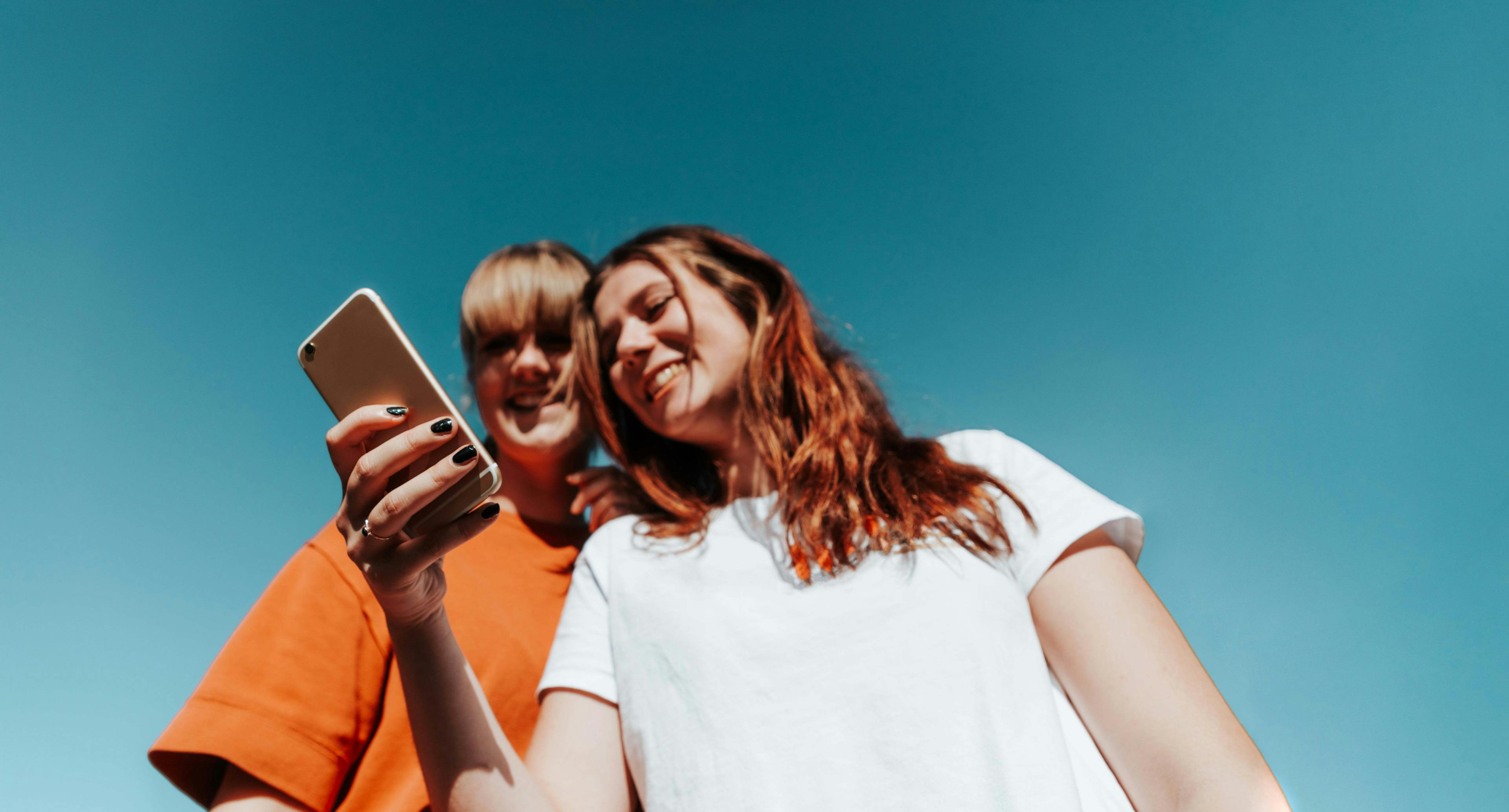 zwei junge Frauen schauen gemeinsam auf ein Smartphone und lachen. Der Hintergrund ist blau.