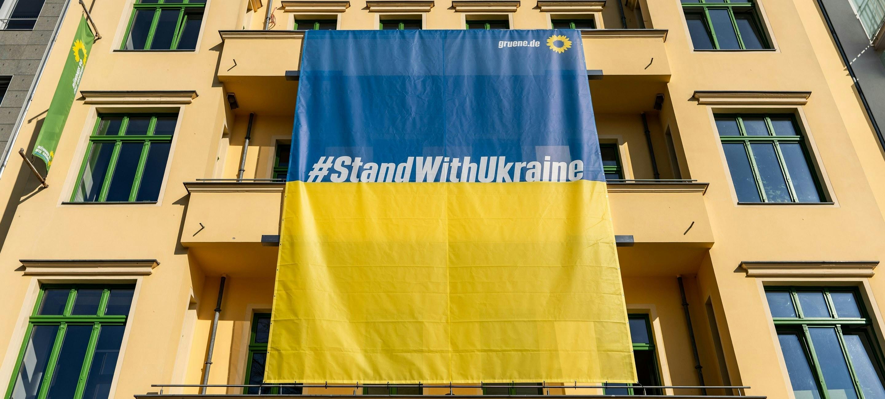 Banner in blau und rot mit der Aufschrift "#StandWithUkraine" hängt an der Fassade eines gelben Hauses.