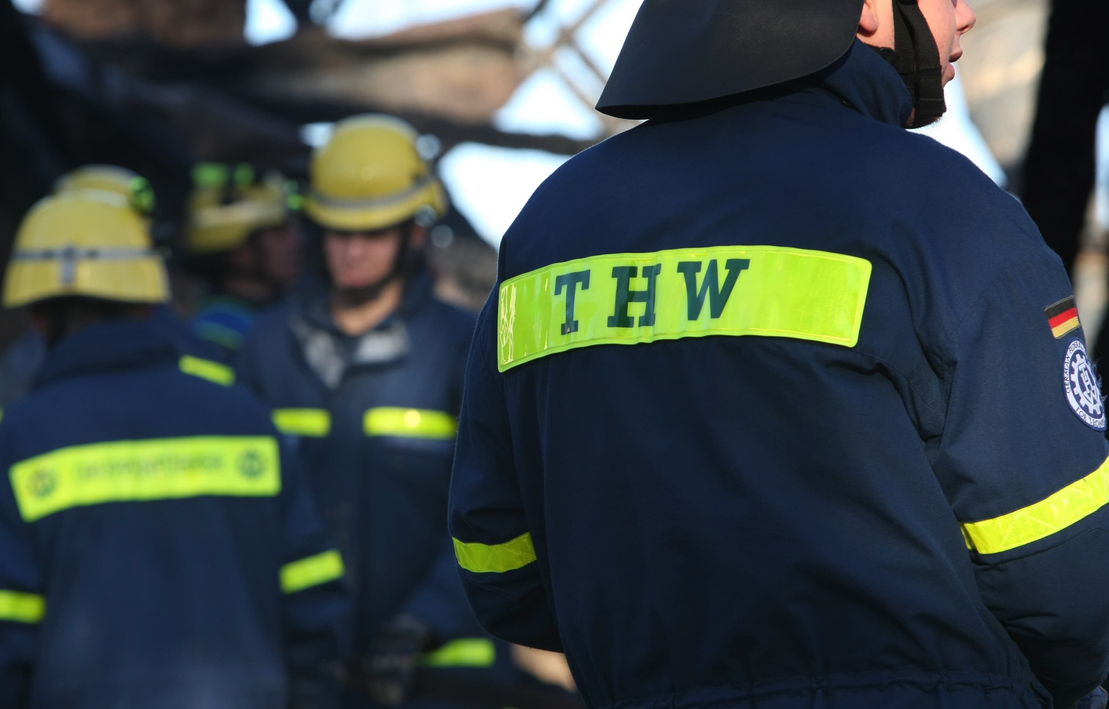 Mann mit dunkelblauer Jacke und einem gelben Streifen mit der Aufschrift "THW" steht mit dem Rücken zur Kamera, im Hintergrund verschwommen zwei weitere Männer mit dunkelblauen Anzügen und gelben Streifen sowie mti gelben Helmen auf dem Kopf.