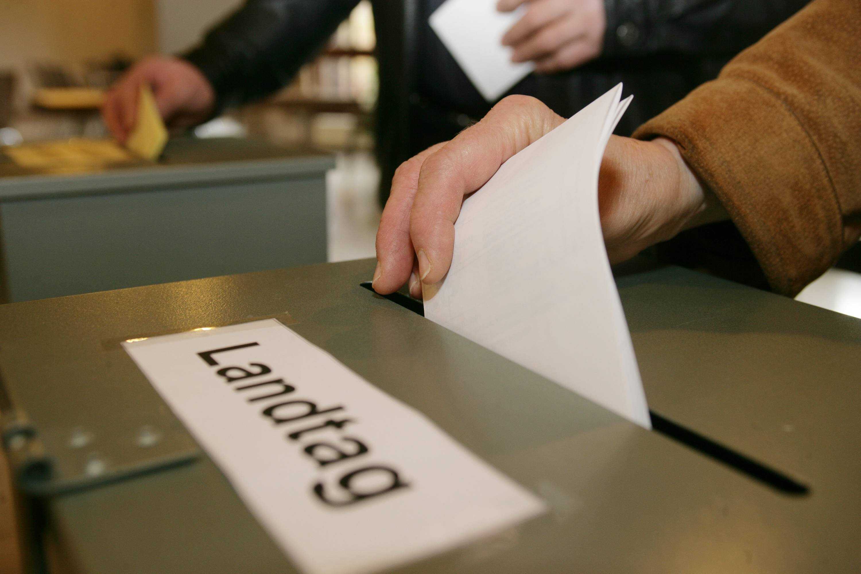 Eine Hand wirft ein weißes, zusammengefaltetes Blatt Papier in einen Behälter mit der Aufschrift "Landtag"