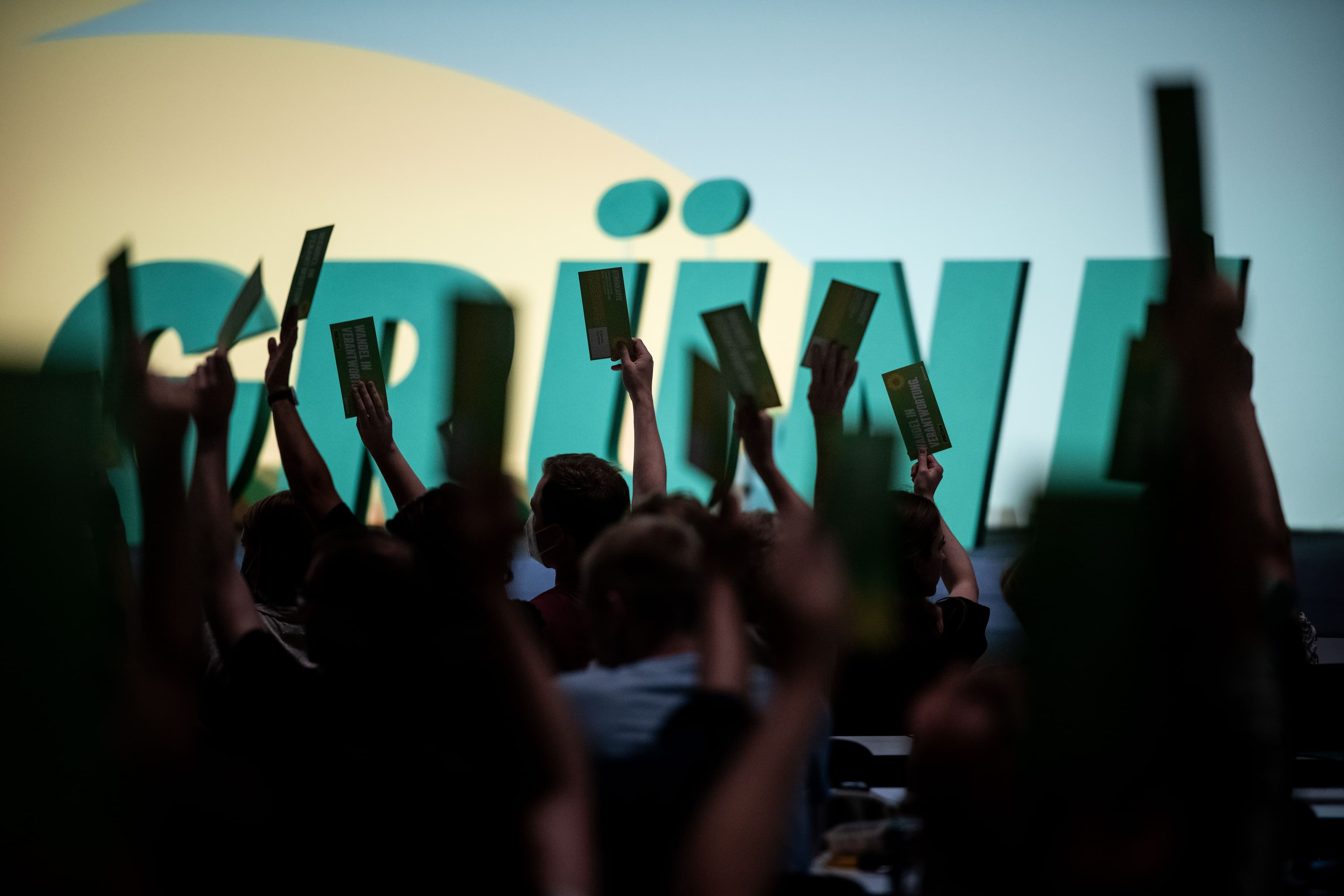 Helle Leinwand ingrün und gelb, vor der große grüne Buchstaben das Wort "GRÜNE" formen. Im Vordergrund sieht man die Silhouetten von vielen Armen, die Zettel in die Höhe strecken.