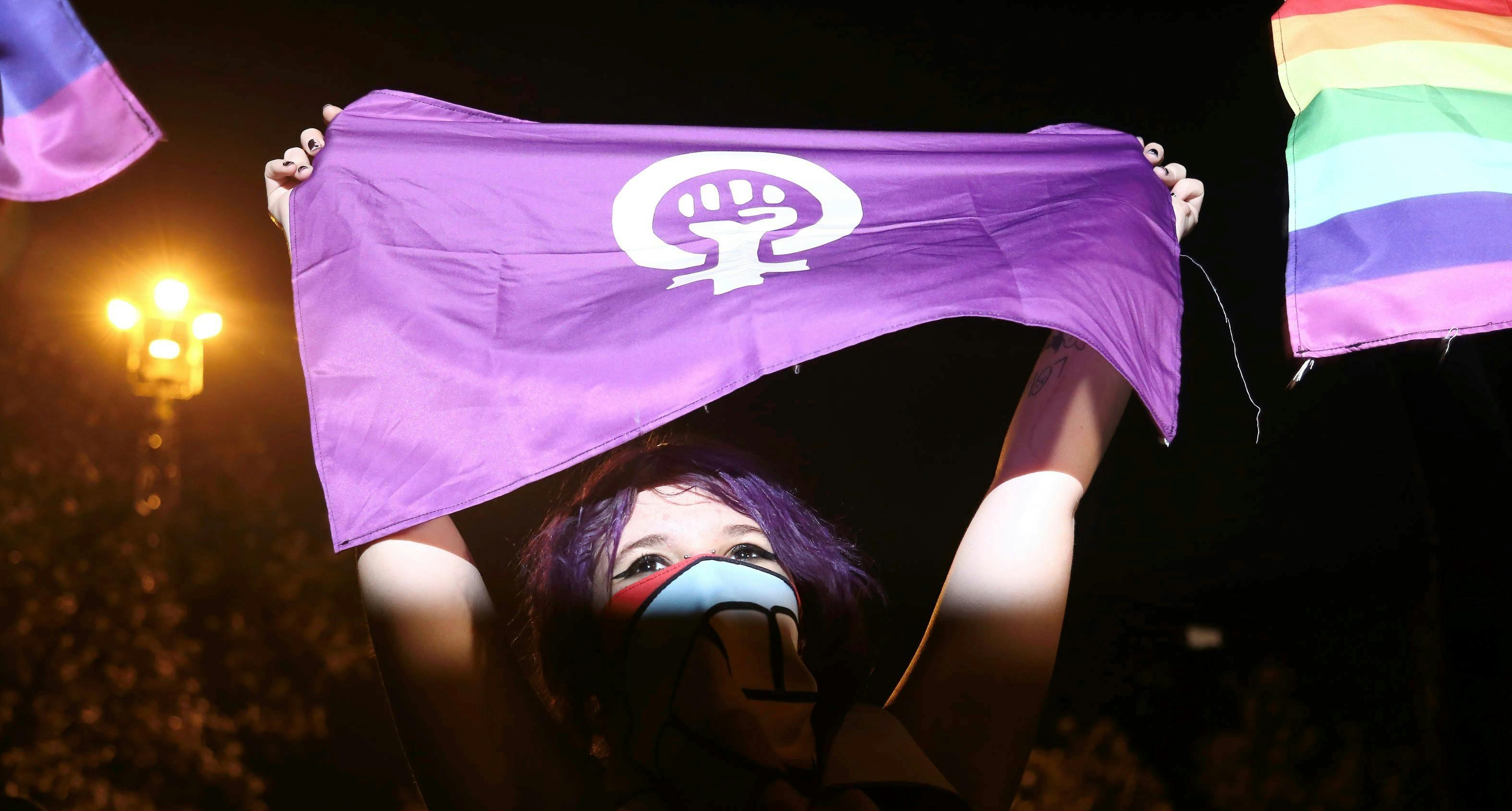 Eine junge Frau steht im Flutlicht. Im Bild sind nur ihr Kopf und ihre Arme. Sie hält eine violette Flagge hoch mit einem weißen Symbol mit einer Faust, die sich in dem Weiblichkeitssymbol befindet. Sie trägt ein Tuch um Mund und Nase.
