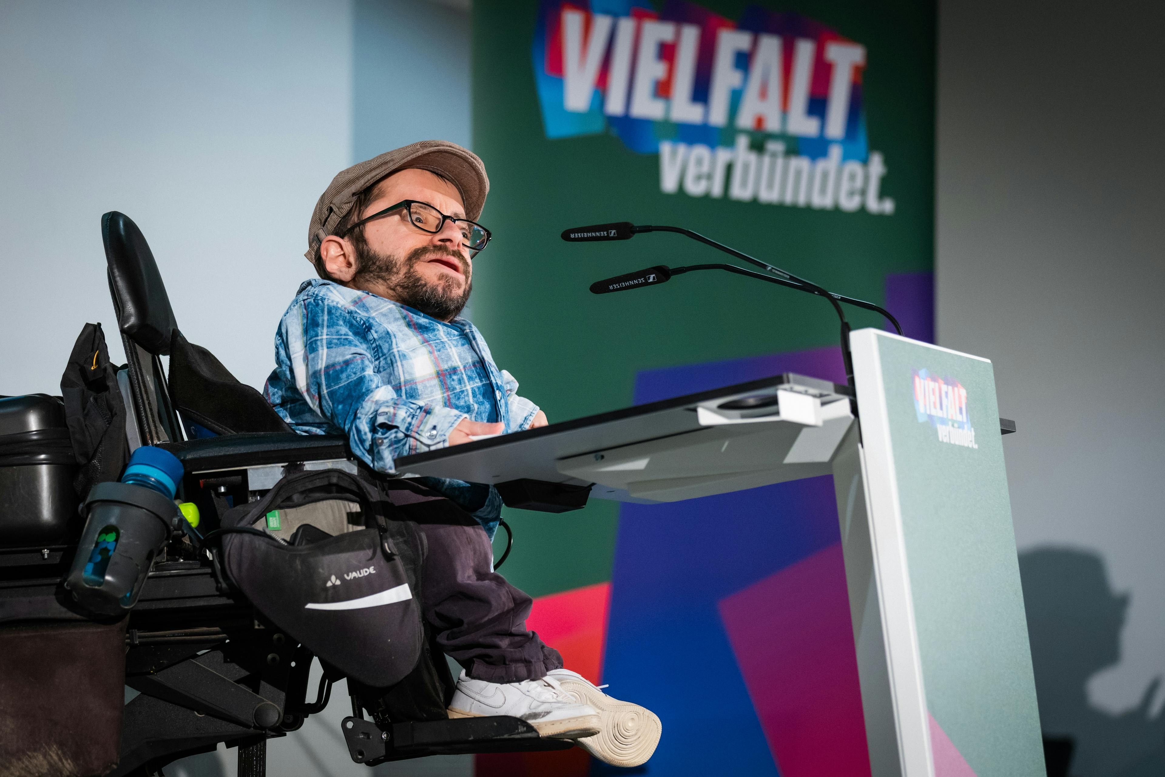Ein Mann im Rollstuhl trägt eine Kappe und spricht an einem Rednerpult in ein Mikrofon. Im Hintergrund befindet sich ein buntes Banner.