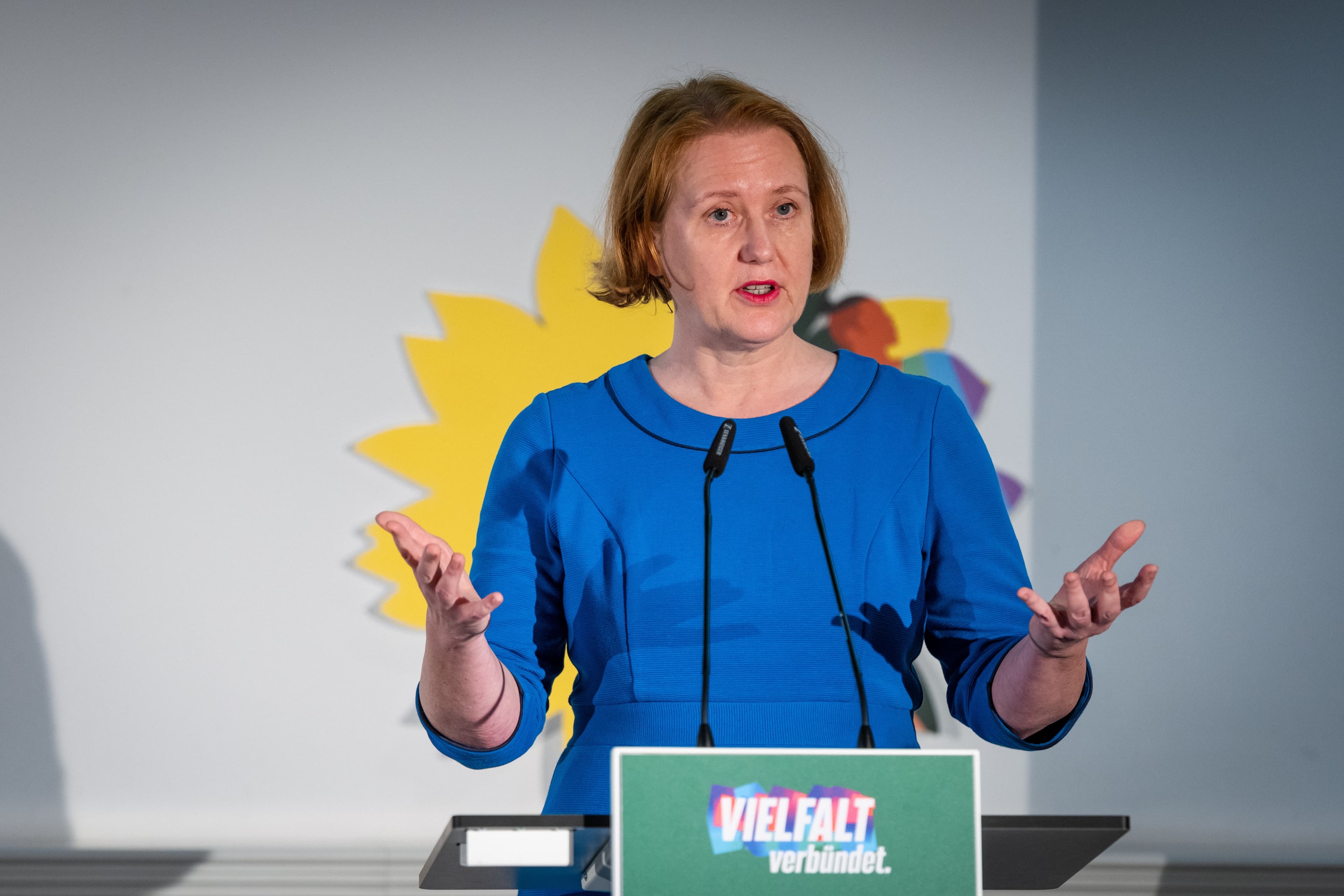 Eine Frau in einem blauen Oberteil spricht an einem grünen Rednerpult. Im Hintergrund befindet sich eine Projektion einer gelben Sonnenblume auf einer weißen Wand.