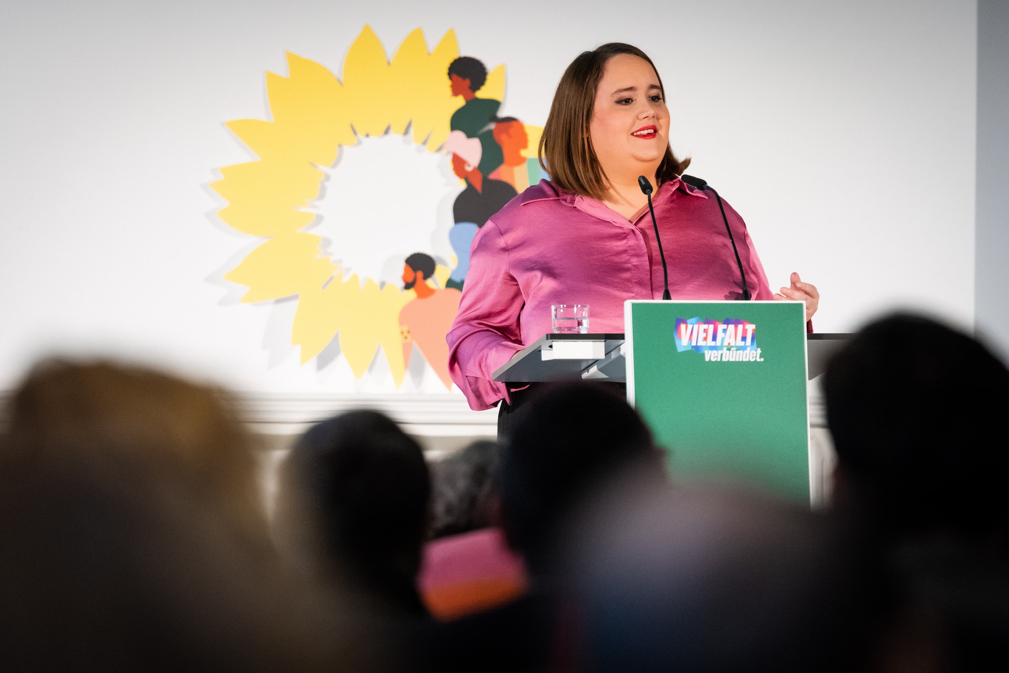 Ricarda Lang steht hinter einem grünen Redner*innenpult mit der Aufschrift "Vielfalt verbündet" und spricht in ein Mikrofon. Im Hintergund sieht man die gelbe Sonneblume mit bunten Grafiken darauf. Im Vordergrund sieht man unscharf die Hinterköpfe von im Publikum sitzenden Menschen.