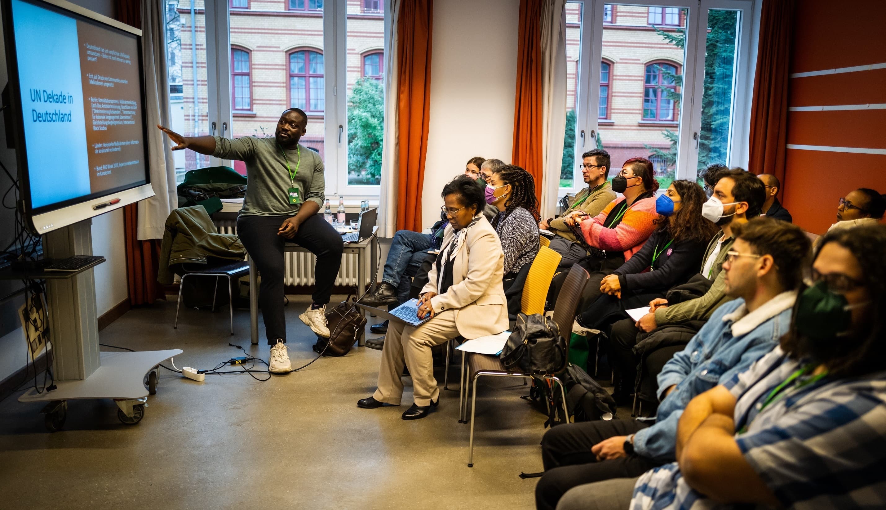 Rechts im Bild sitzt eine Gruppe Menschen auf Stühlen und hört einem Mann links im Bild zu. Alle schauen auf eine Leinwand.