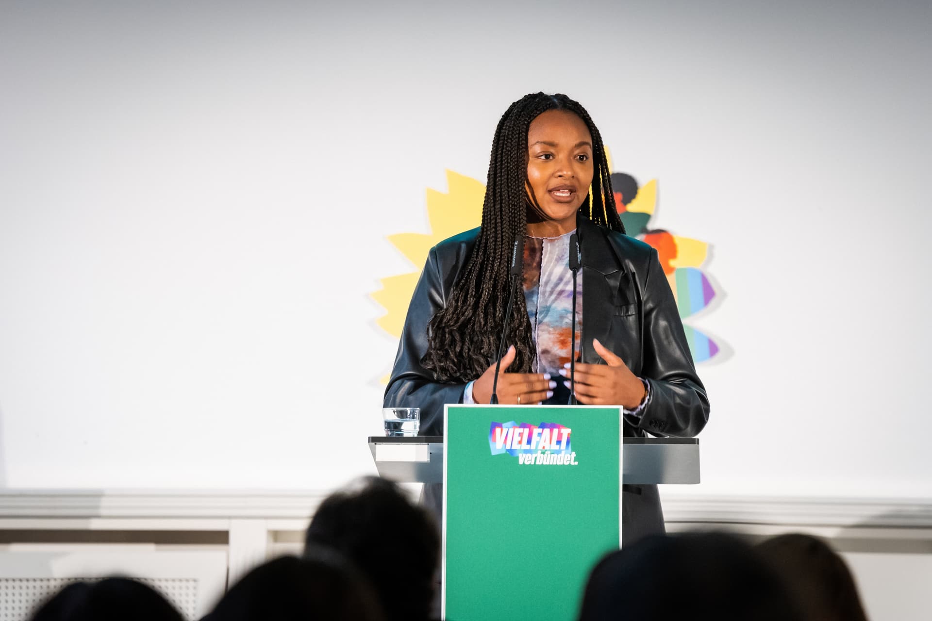 Aminata Touré steht hinter einem grünen Redner*innenpult mit der Aufschrift "Vielfalt verbündet" und spricht in ein Mikrofon. Im Hintergund sieht man die gelbe Sonneblume mit bunten Grafiken darauf. Im Vordergrund sieht man unscharf die Hinterköpfe von im Publikum sitzenden Menschen.