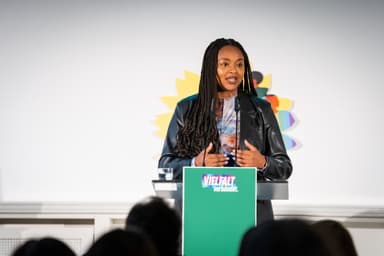 Aminata Touré steht hinter einem grünen Redner*innenpult mit der Aufschrift "Vielfalt verbündet" und spricht in ein Mikrofon. Im Hintergund sieht man die gelbe Sonneblume mit bunten Grafiken darauf. Im Vordergrund sieht man unscharf die Hinterköpfe von im Publikum sitzenden Menschen.