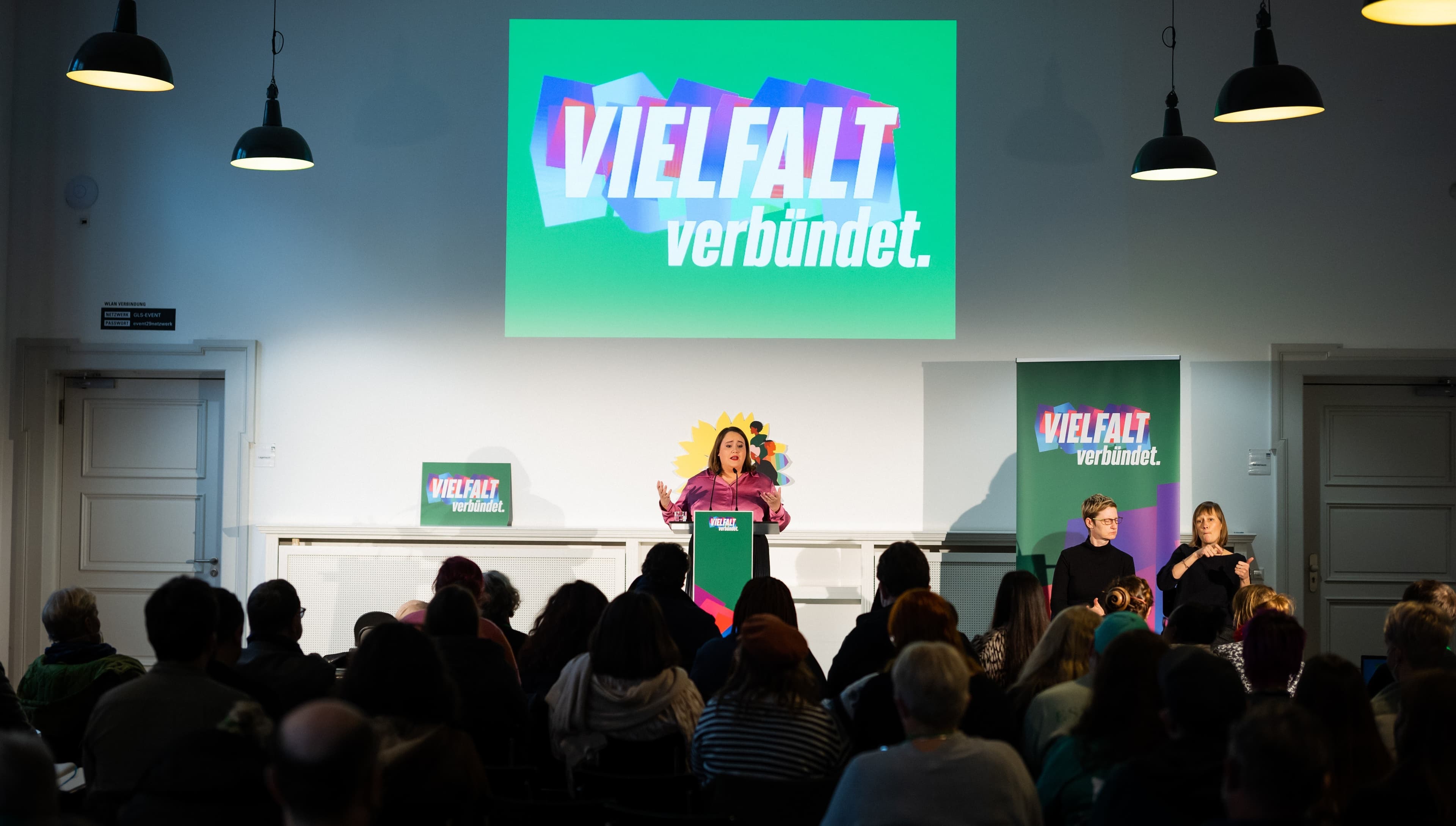 Totale mit dem Publikum von hinten, das auf die Bühne schaut, auf der Ricarda Lang hinter einem Redner*innenpult steht. Über ihr hängt ein grünes Transparent mit der Aufschrift: "Vielfalt verbündet"