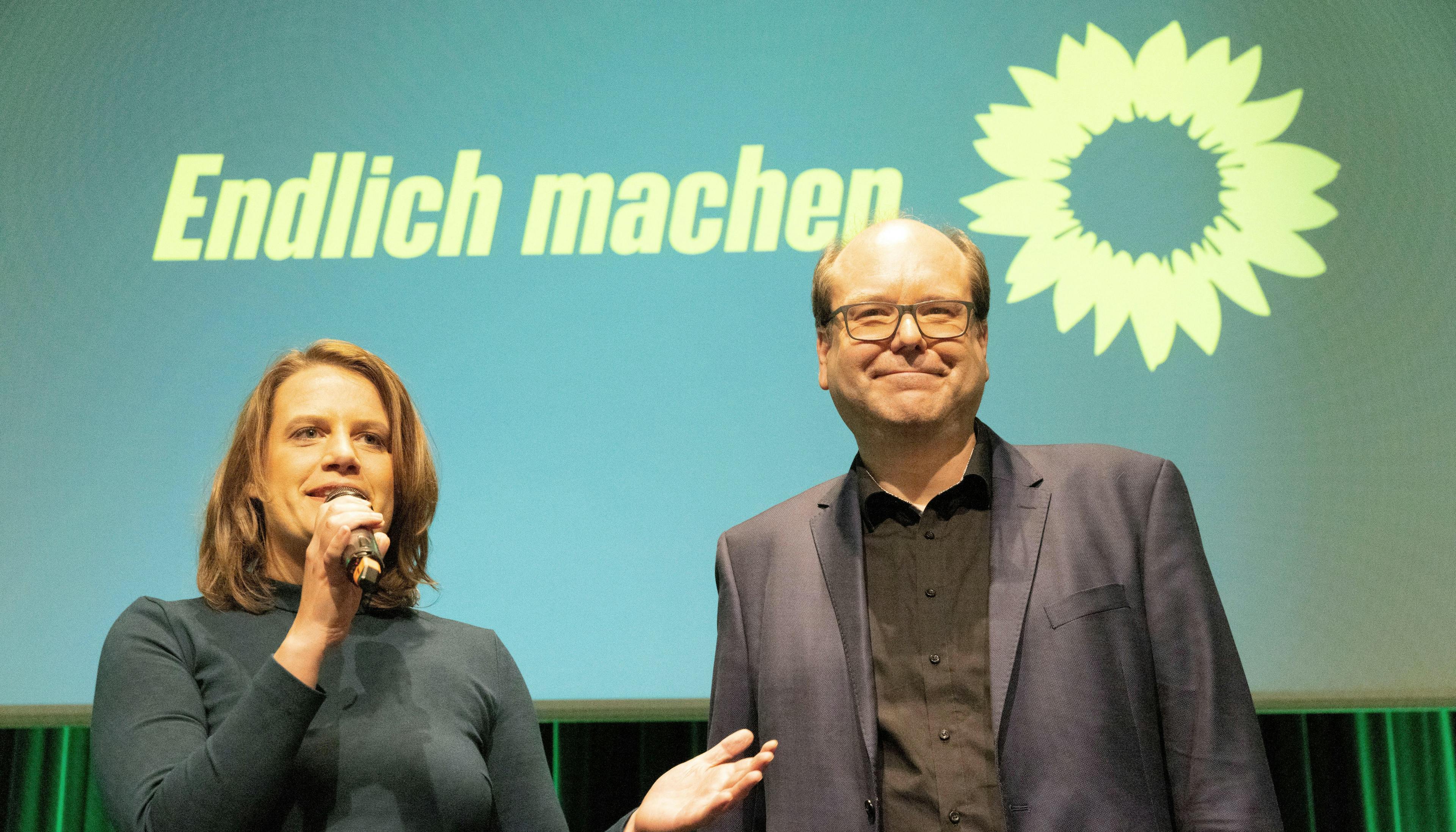 Das grüne Spitzenduo Julia Willie Hamburg und Christian Meyer steht vor einer Leinwand, auf der das Sonnenblumen-Logo abgebildet ist und in gelber Schrift steht: "Endlich machen". Julia Willie Hamburg spricht in ein Mikrofon.
