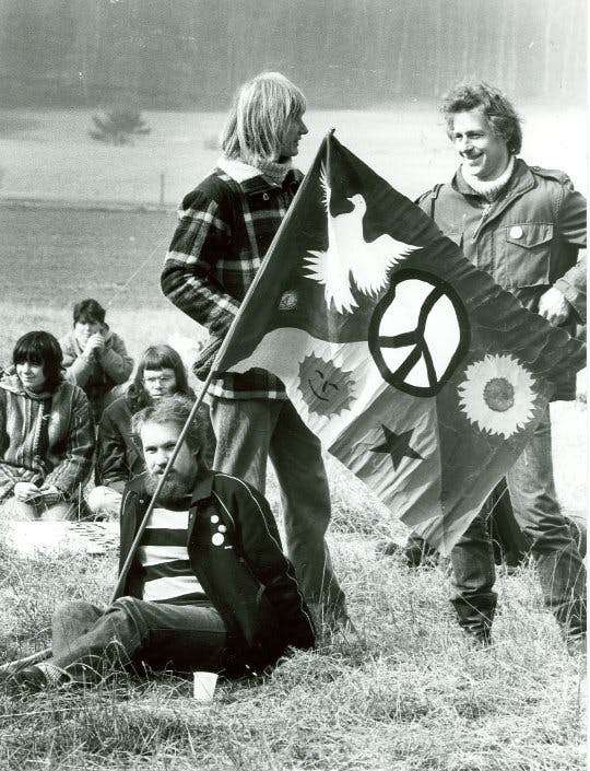 Schwarz-weiß Foto: Mehrere Menschen mit langen Haaren sitzen auf einer Wiese. Im Vordergrund ist eine Fahne mit Friedenszeichen, Sonnenblume, Stern und lachender Sonne zu sehen.