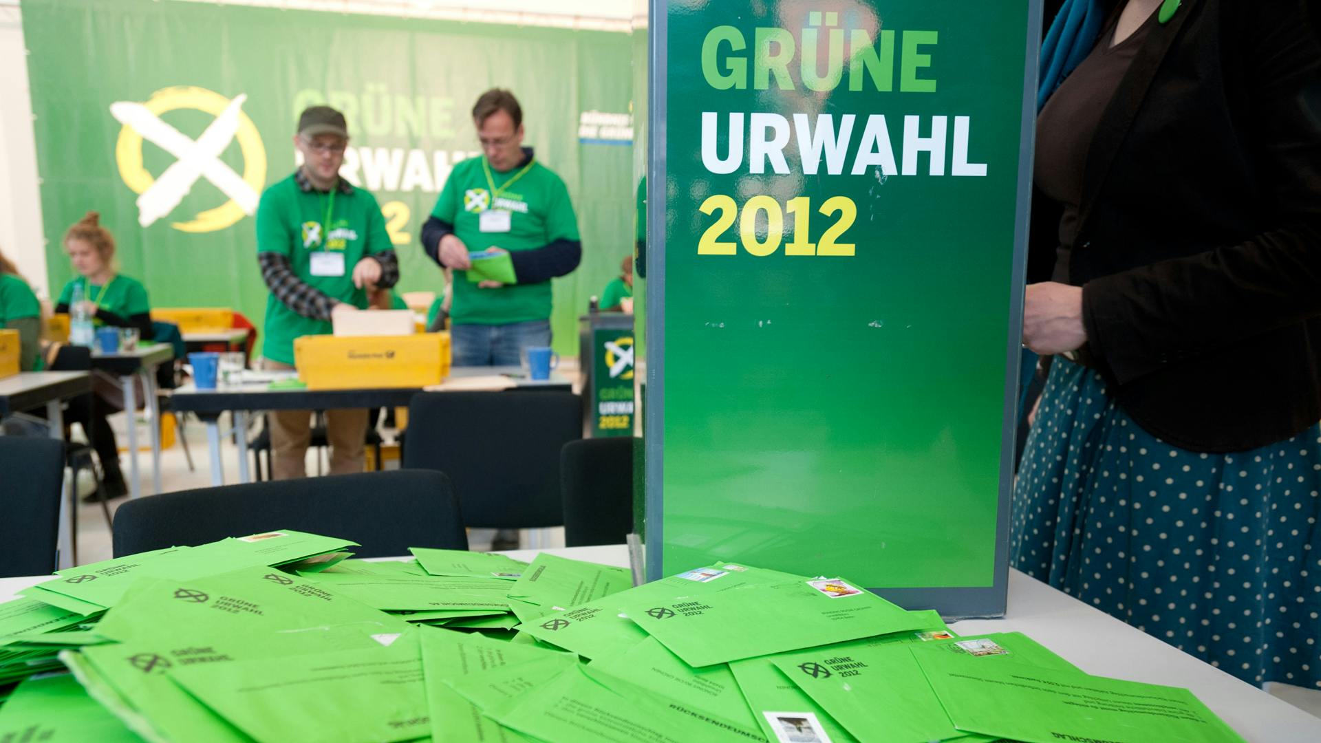 Im Vordergrund liegen viele grüne Briefumschläge auf einem Tisch. Dahinter ein Aufsteller, auf dem "Grüne Urwahl 2012" steht sowie mehrere Menschen, die Briefe aus Postkisten nehmen und öffnen.