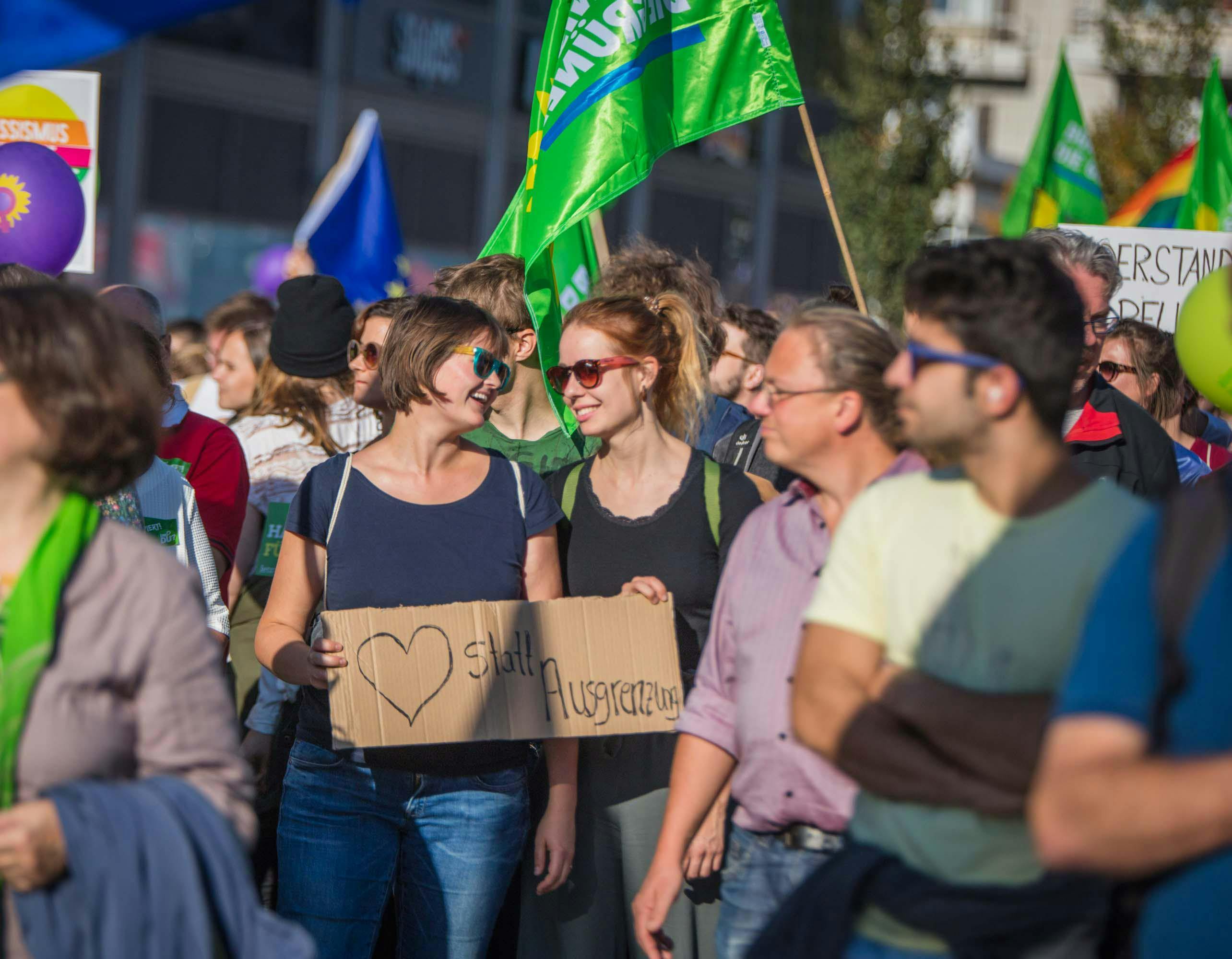 Grüne Mitglieder demonstrieren in Berlin und halten dabei Schilder und Fahnen hoch. Im Bildmittelpunkt stehen zwei junge Frauen, die sich anlächeln.