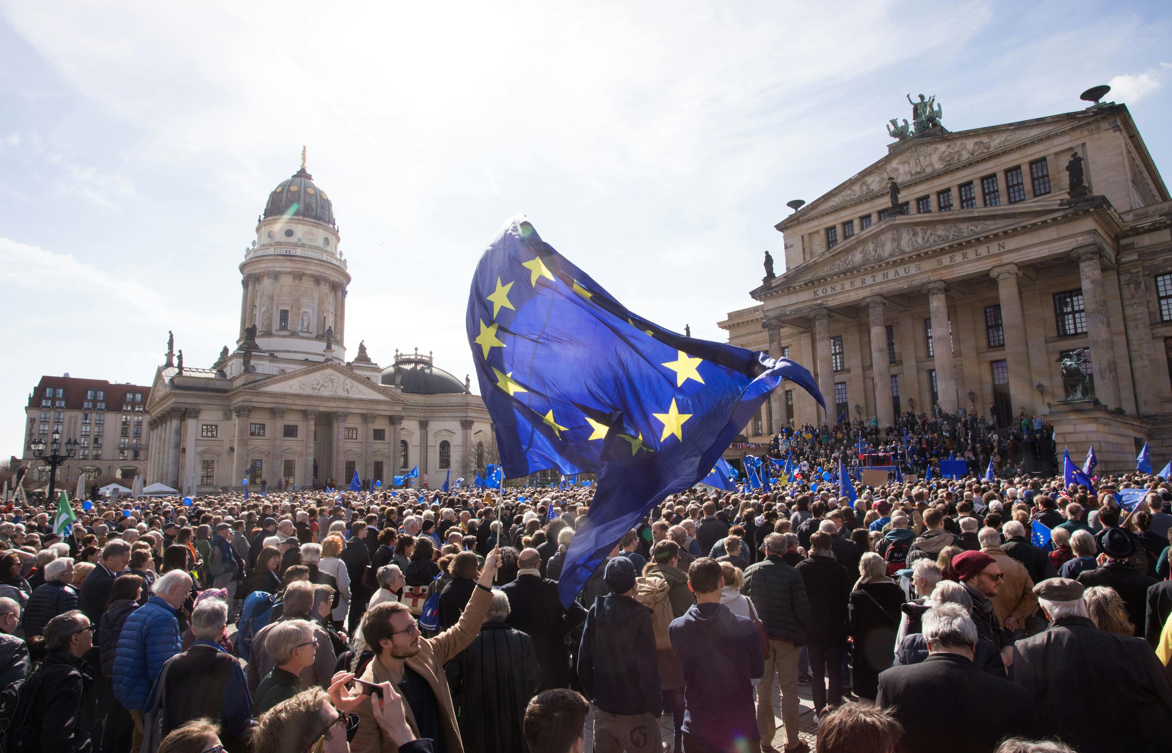 Bei einer Pulse of Europe Demo auf dem Gendarmenmarkt in Berlin stehen vor der schöner Kulisse der Dome viele Menschen und feiern Europa. Zentral im Fokus sieht man eine große Europaflagge, die jemand schwingt.