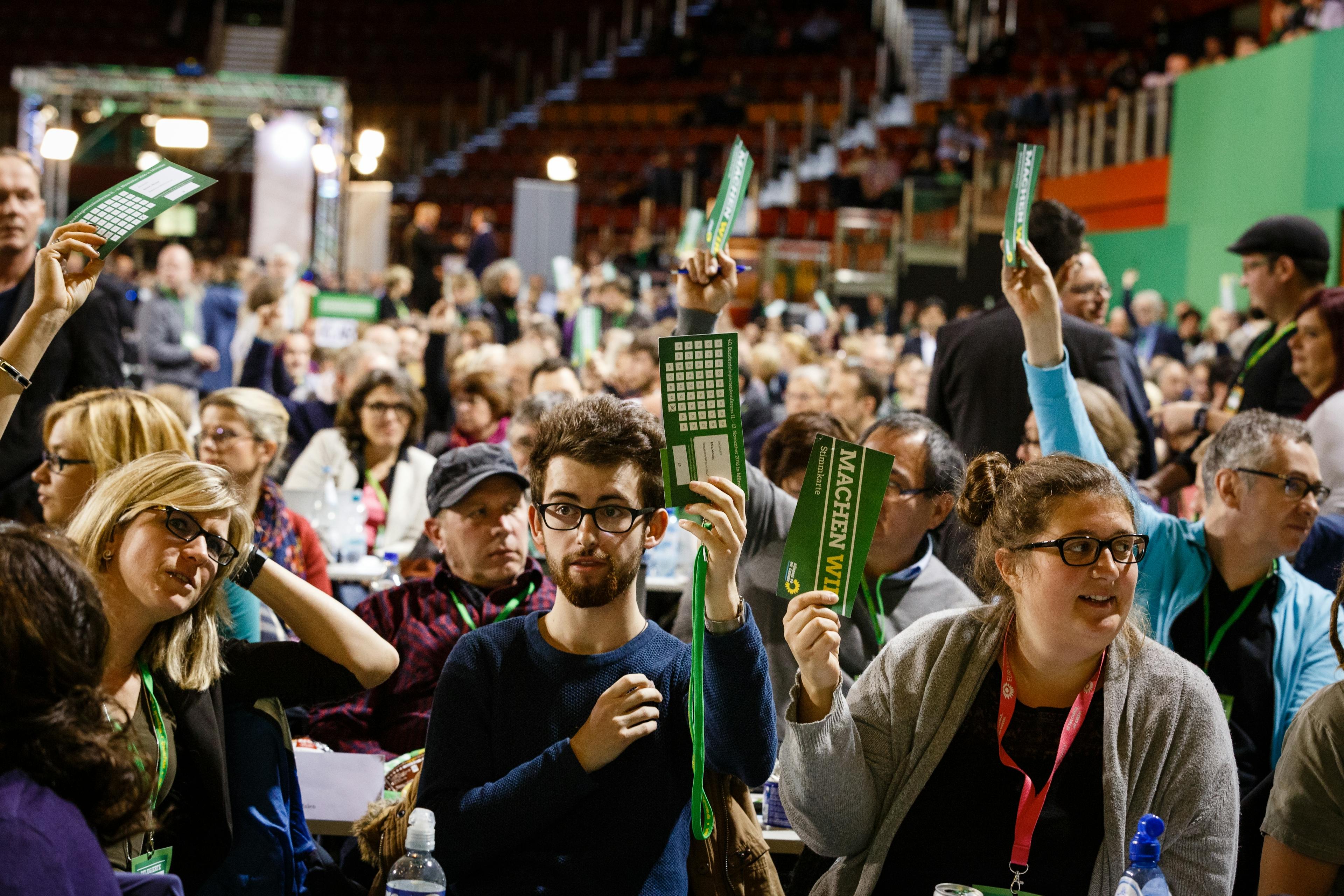Grüne Parteimitglieder setzen in einem großen Saal während des Parteitages und halten Stimmzettel in die Höhe.