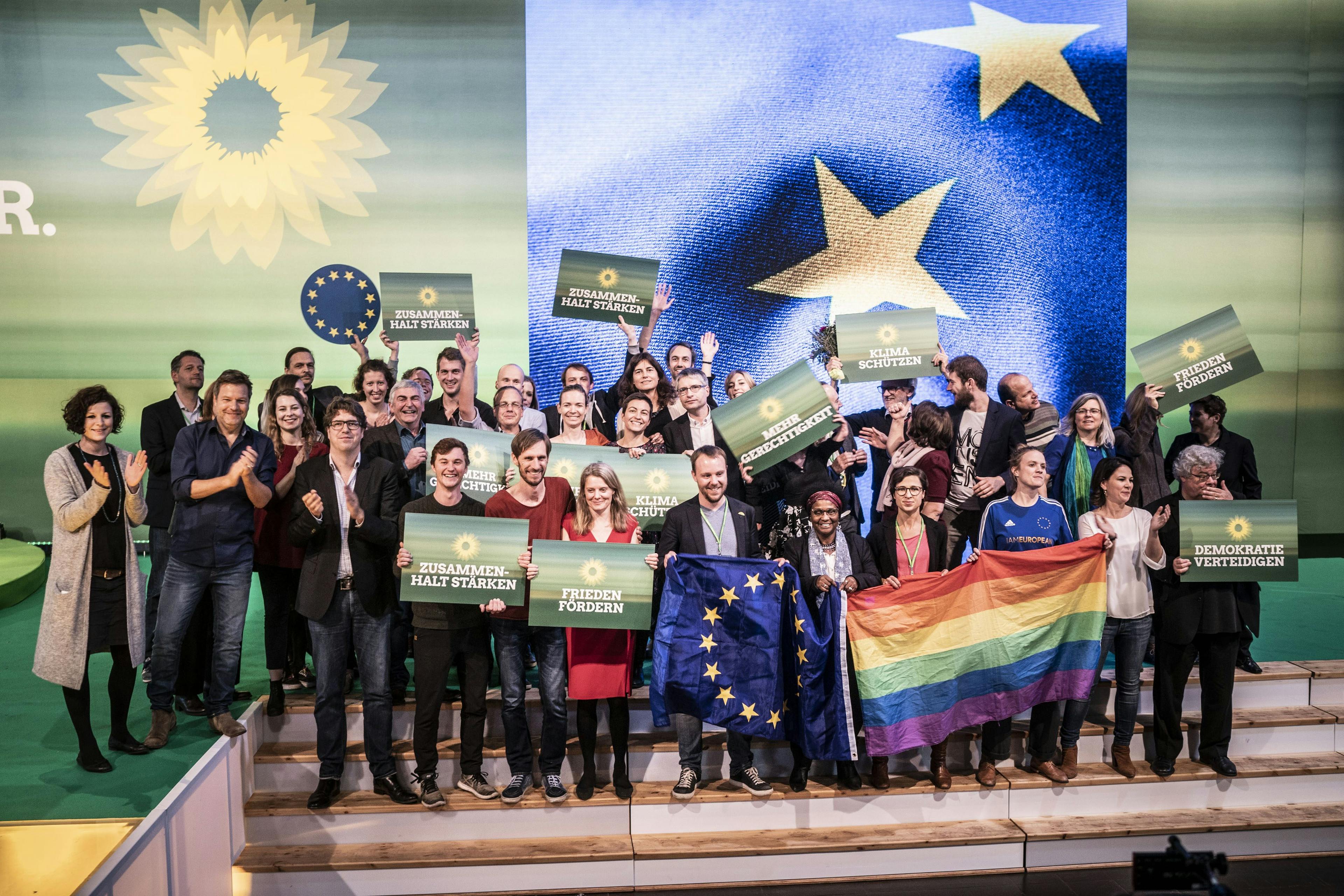 Mehrere Personen stehen auf dem Treppenabsatz einer Bühne und halten Schilder in die Höhe. Im Hintergrund ist ein Ausschnitt der Europaflagge zu sehen.